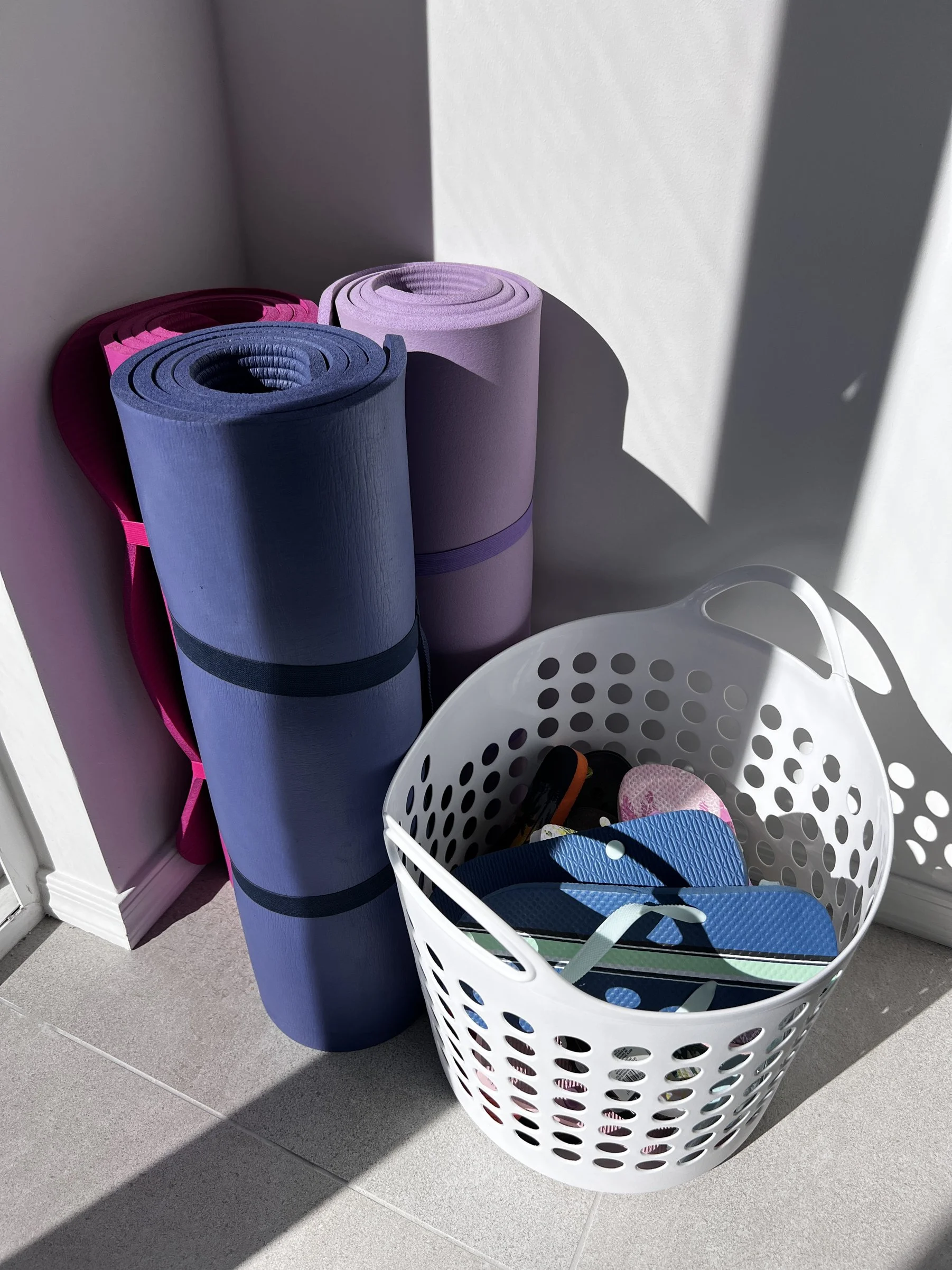 Three rolled-up yoga mats, one pink, one purple, and one blue, standing upright next to a white plastic basket filled with colorful flip-flops and sandals, on a tiled floor in a sunlit area.
