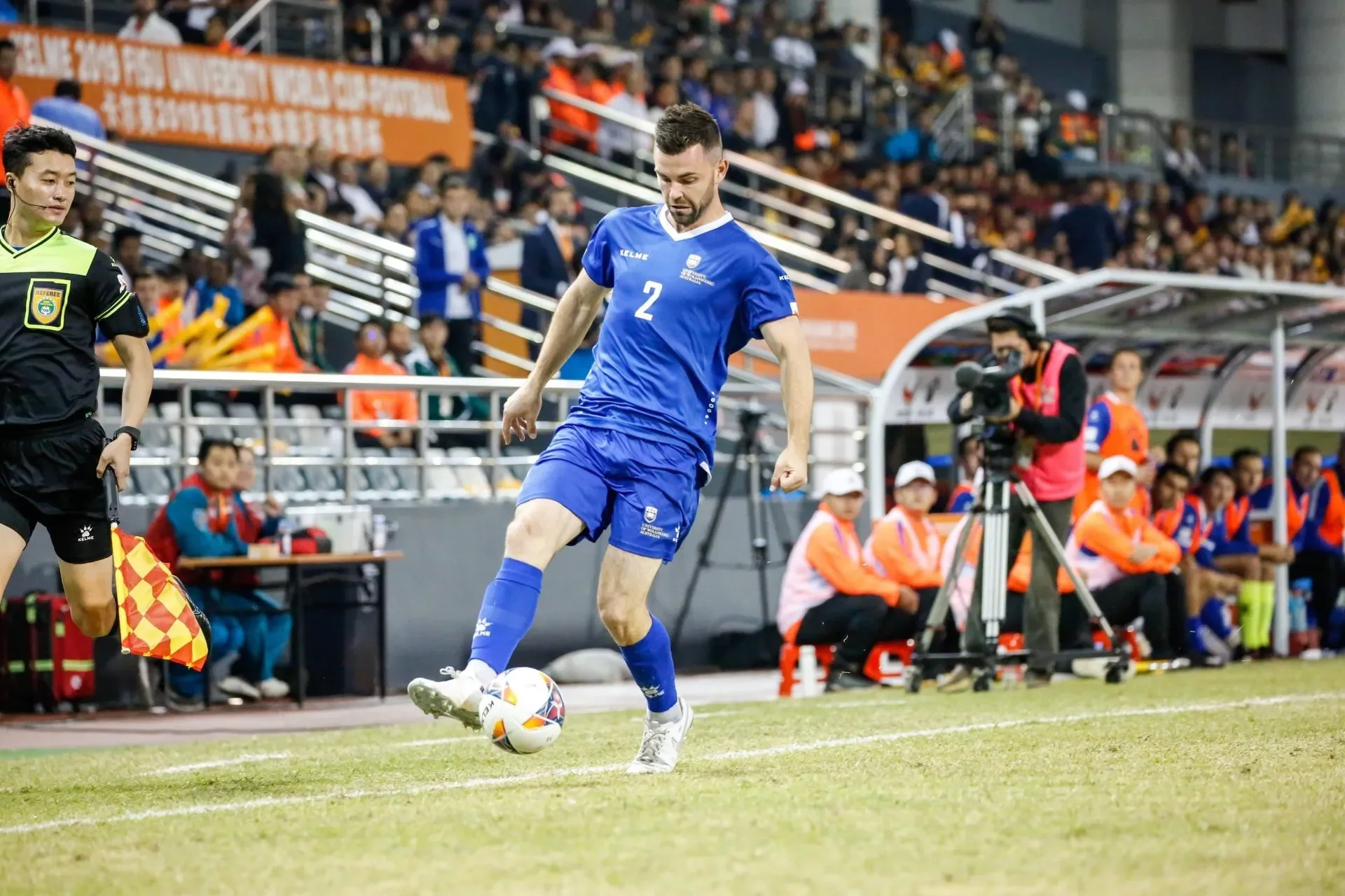 A soccer player in a blue uniform with the number 2, kicking a soccer ball during a match. An assistant referee is nearby, and the sidelines show substitutes, coaches, and officials. Spectators fill the stands in the background.