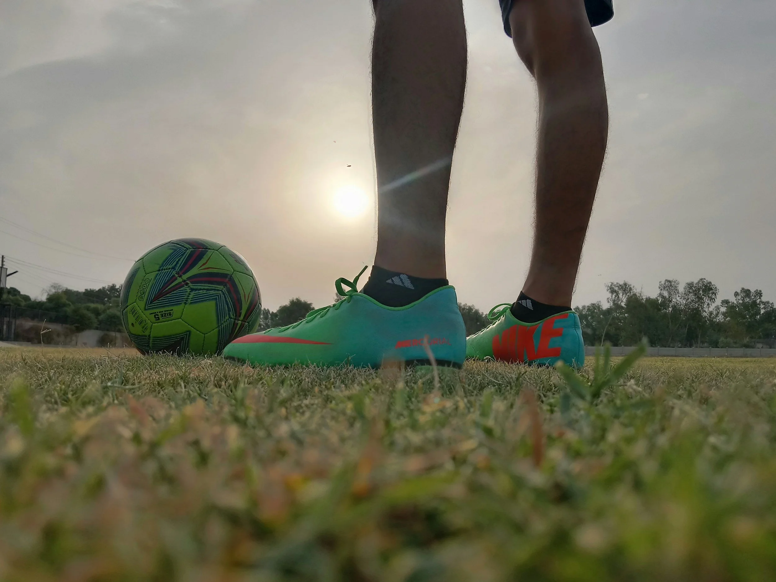 Close-up of a person's legs and feet on a soccer field, wearing Nike soccer shoes, with a brightly colored soccer ball next to their feet. The sun is setting or rising in the background.