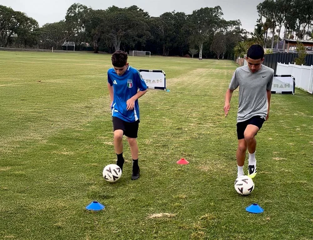 Two boys practicing soccer on a grassy field with cones and small goals.