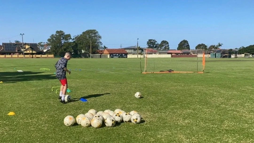 A young boy preparing for soccer practice on a grassy field with a pile of soccer balls, agility cones, and a small soccer goal in the background.