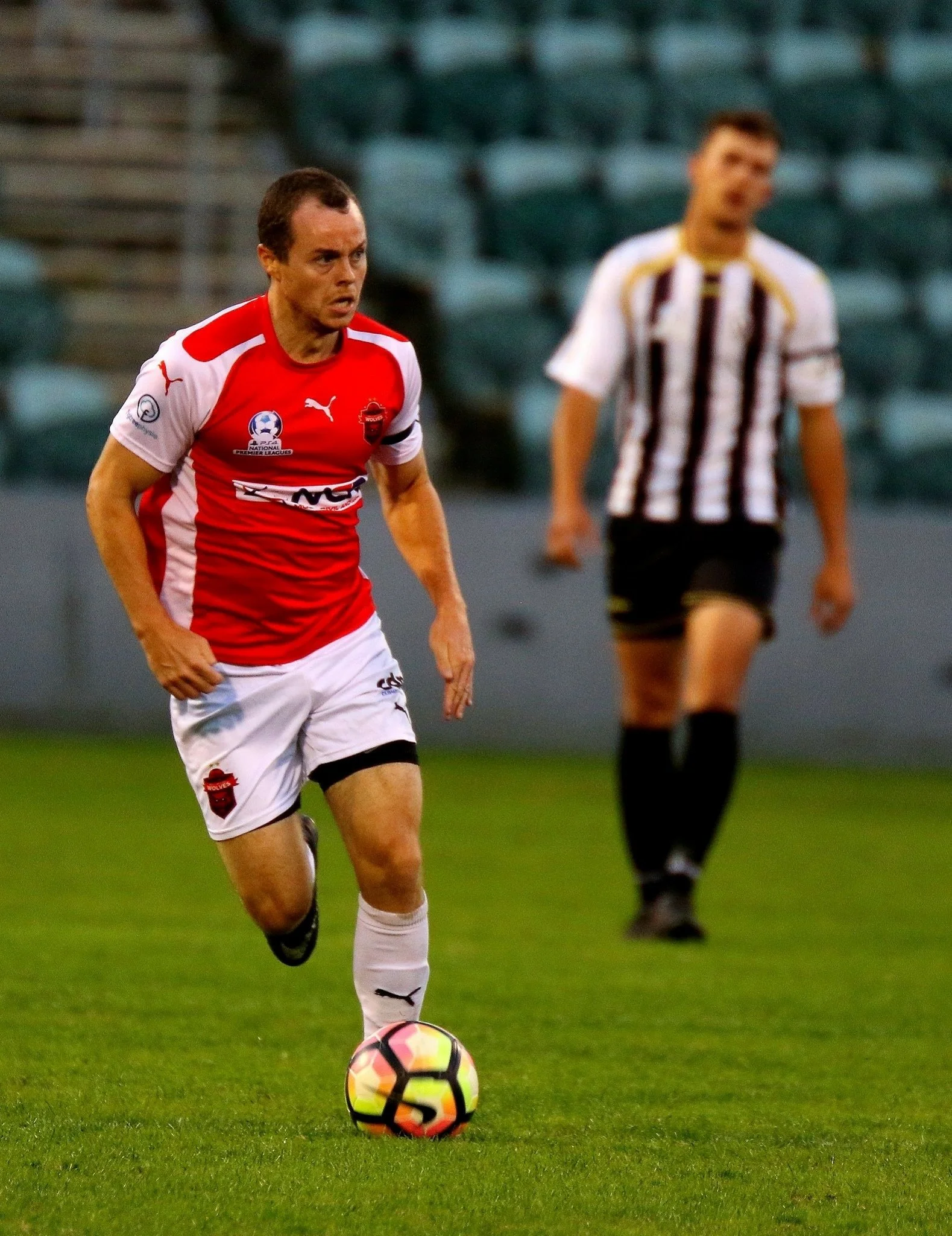 A soccer player in a red and white jersey is running with a soccer ball on a field, with another player in a black and white striped jersey in the background.
