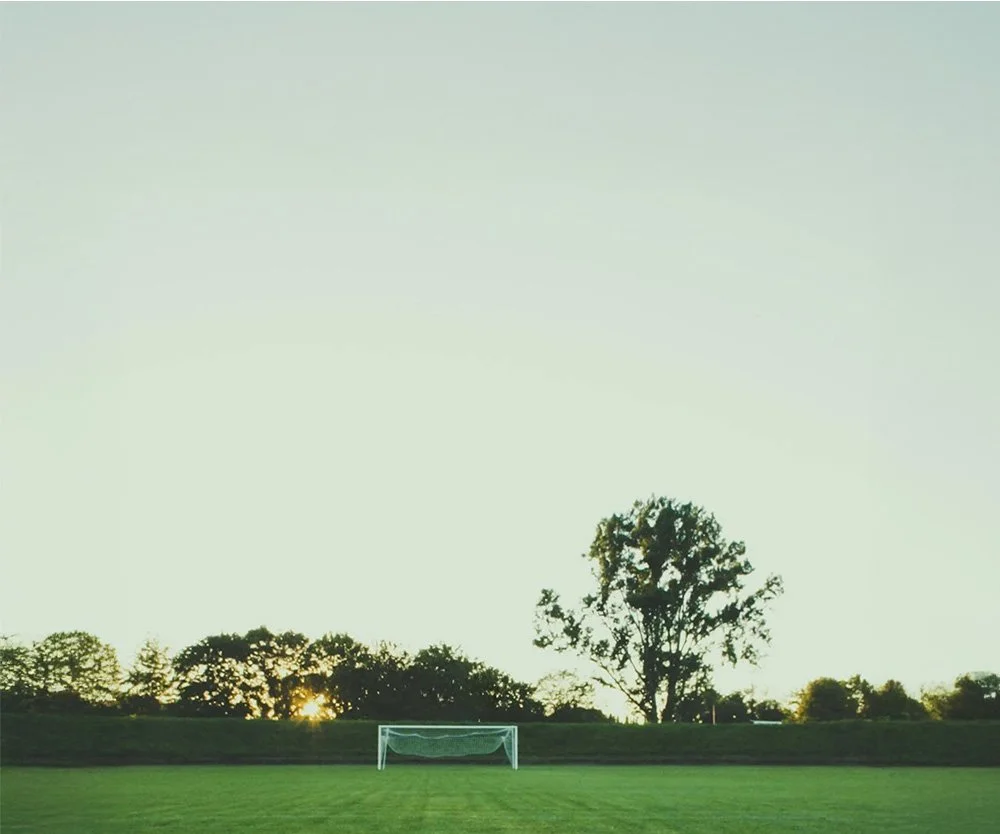 A soccer field with goalpost, trees, and setting sun in the background.