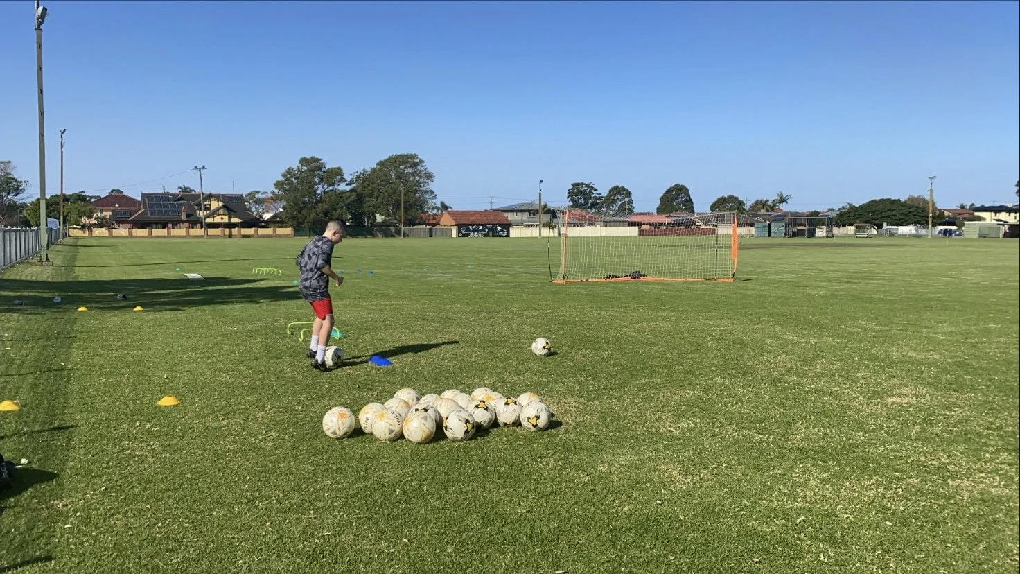 A young boy in a grey and black camouflage shirt and red shorts preparing to kick a soccer ball on a grassy field. There are multiple soccer balls clustered together on the ground, orange cones, a small goal, and training equipment such as hurdles and ladders scattered around. Residential houses are visible in the background under a clear blue sky.