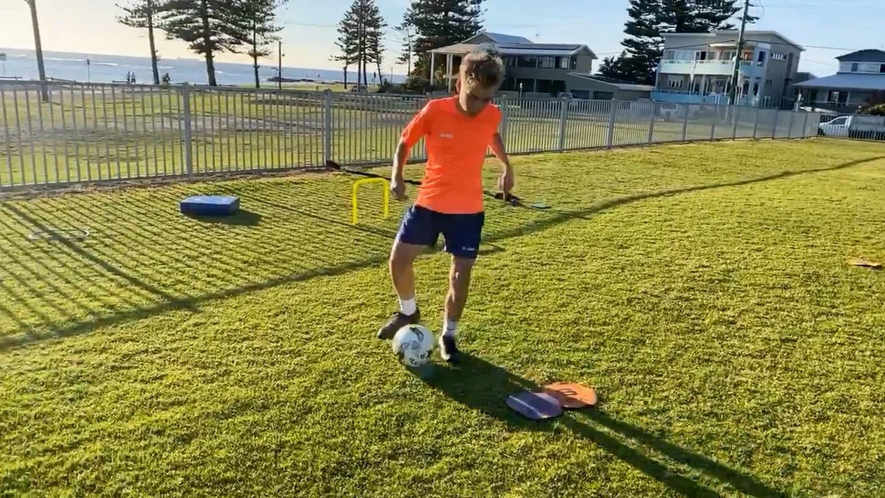 A boy playing soccer on a grassy field near houses, with a ball at his feet and training equipment nearby.