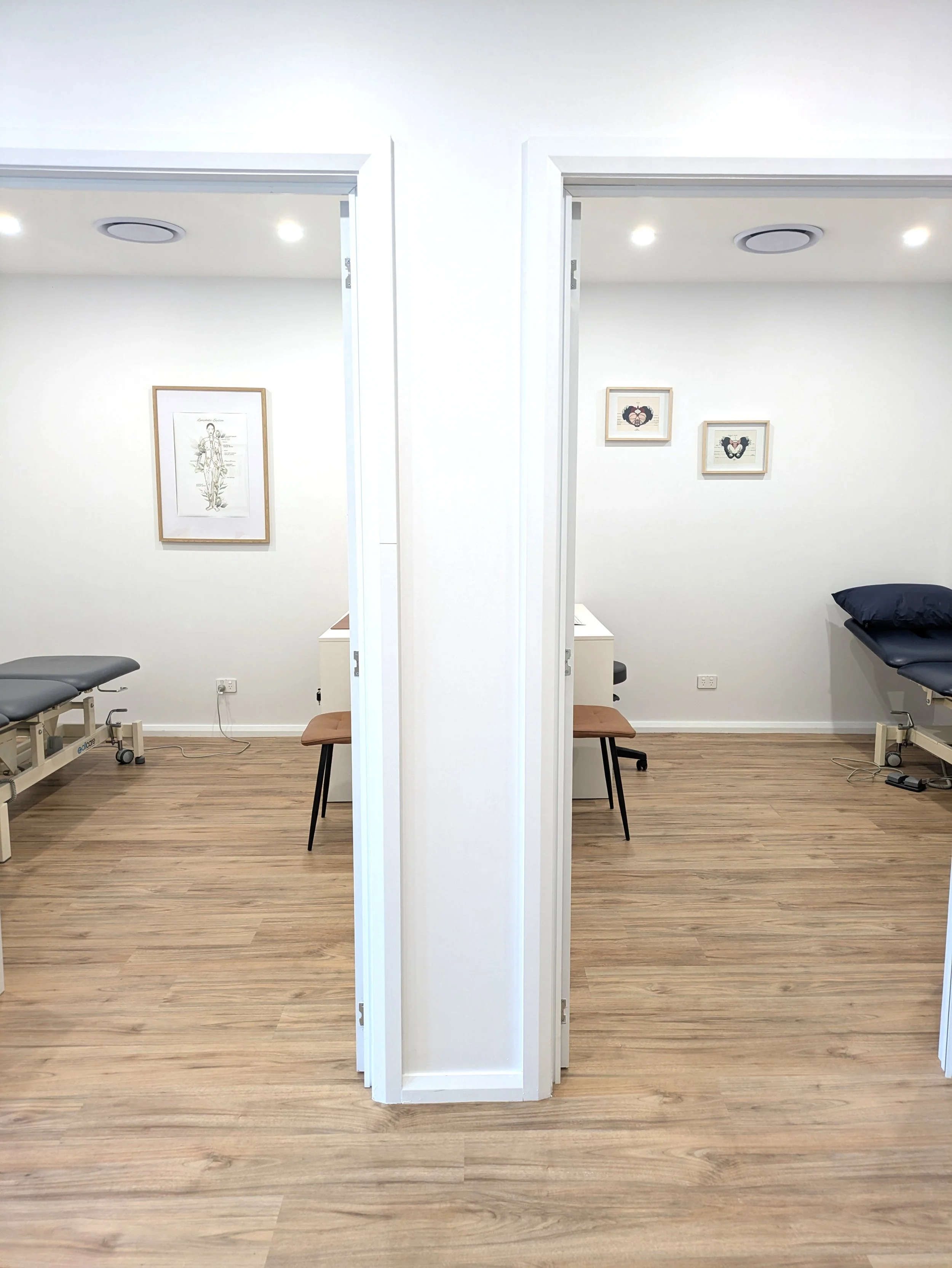 Two examination rooms with white walls, wood flooring, framed artwork, and exercise tables, separated by a wall.