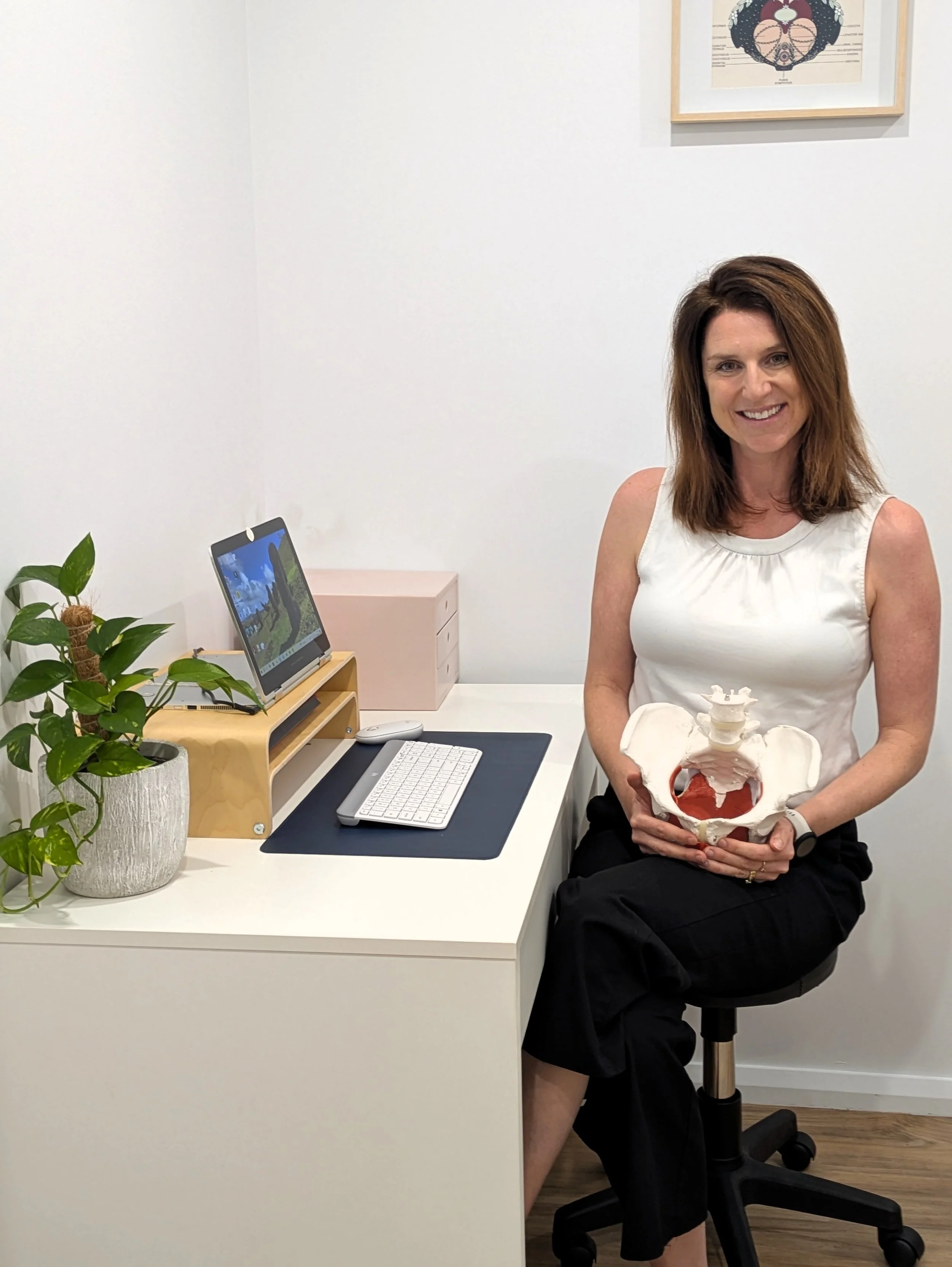 Woman smiling seated at desk holding a pelvis model in a medical or therapy office.