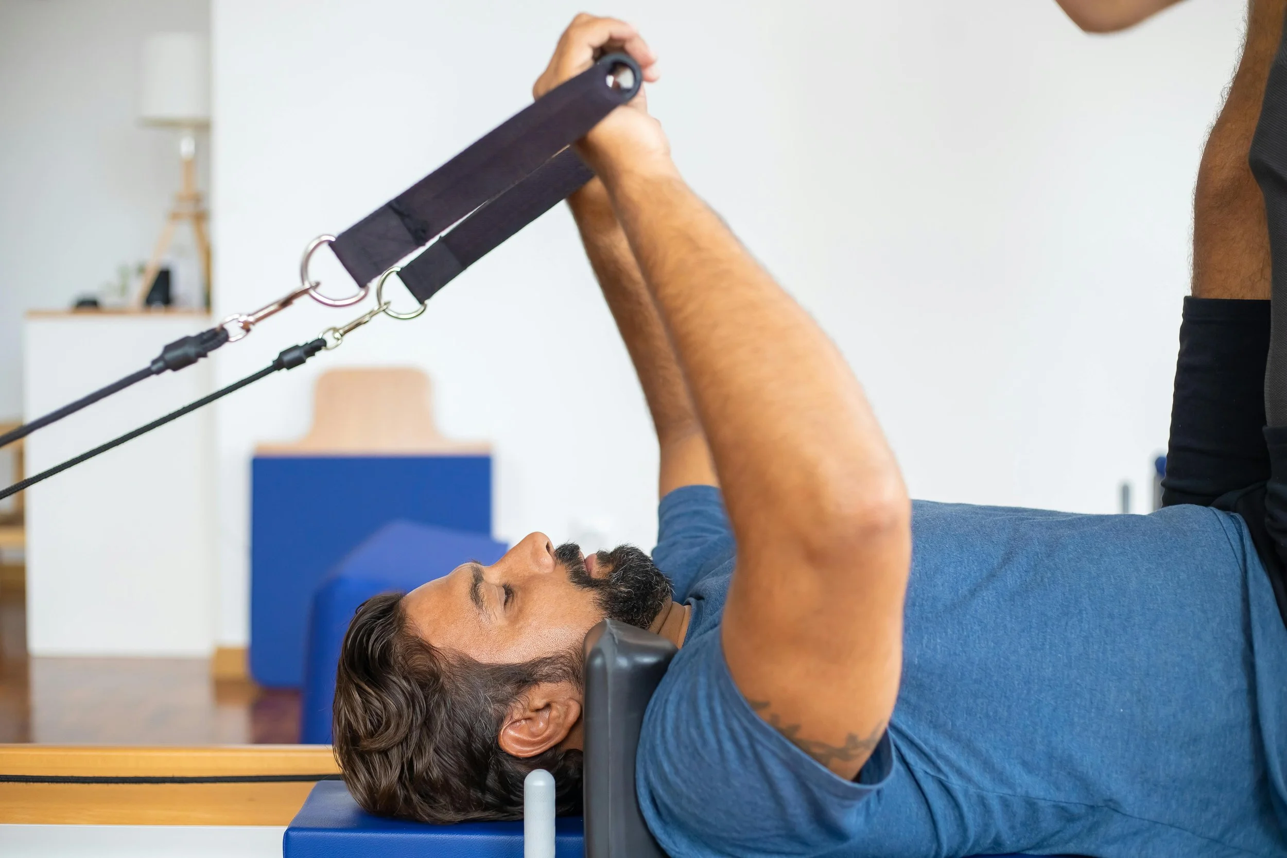 Man lying on a bench while doing a resistance exercise with a cable machine, with a trainer assisting.