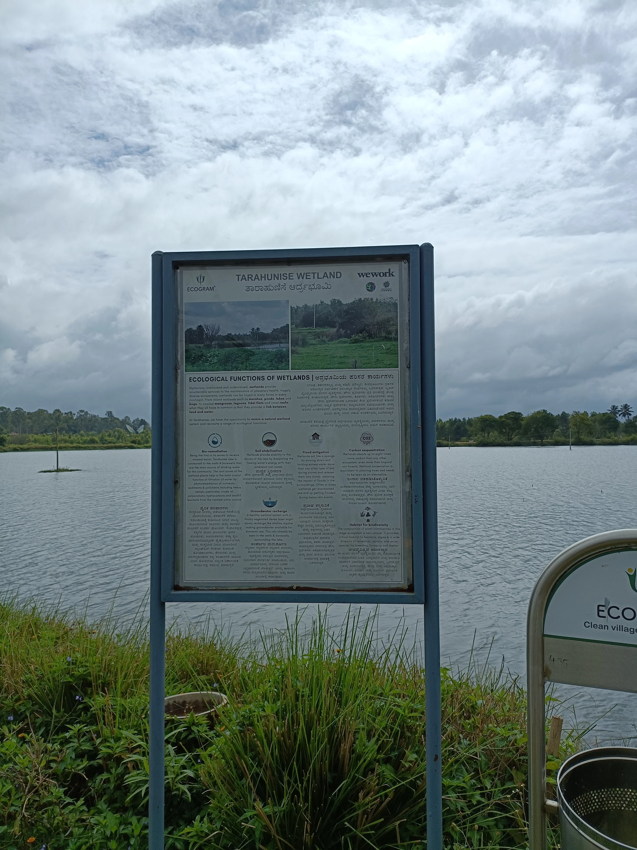 Information signboard at Tarahunise Wetland with a lake and cloudy sky in the background.