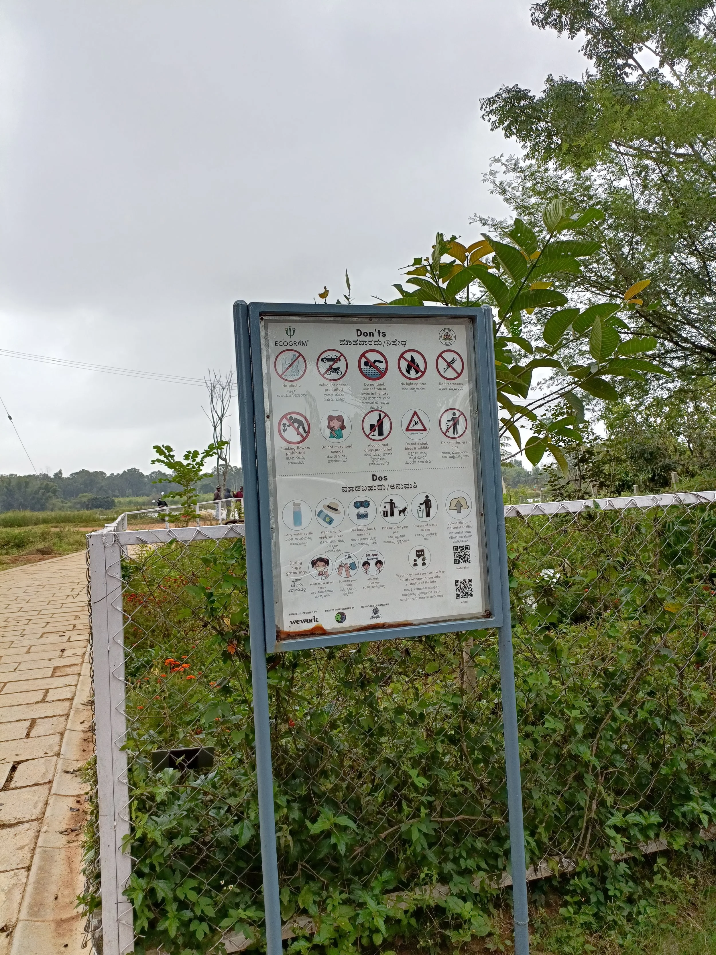 A signboard outdoors with safety and conduct rules in English and another language, displayed on a stand, surrounded by green foliage and a fence, under a cloudy sky.