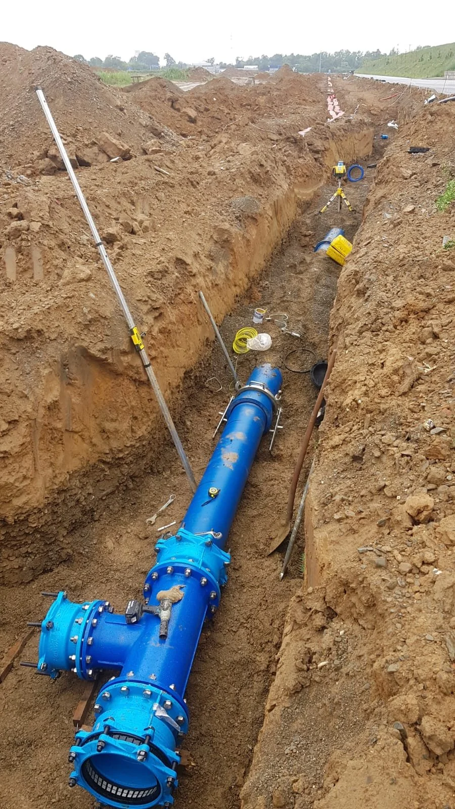 Construction site with a large blue pipeline installed in a trench, surrounded by soil and construction tools.