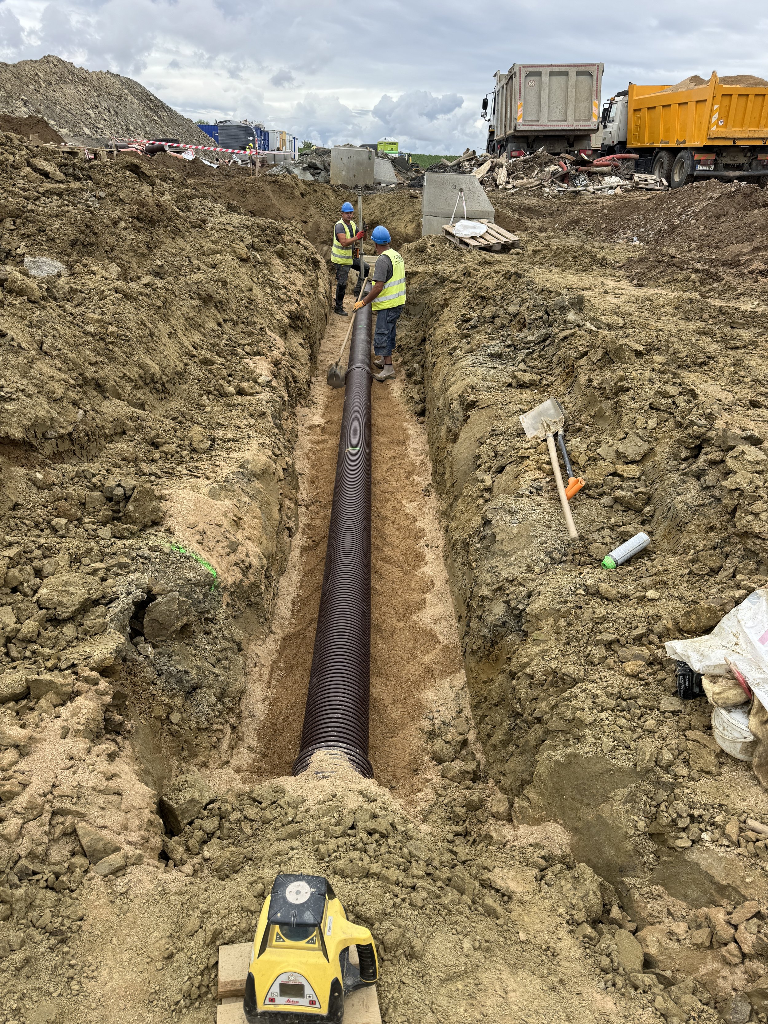 Construction workers installing large underground pipe in a trench at a construction site with trucks and gravel in the background.