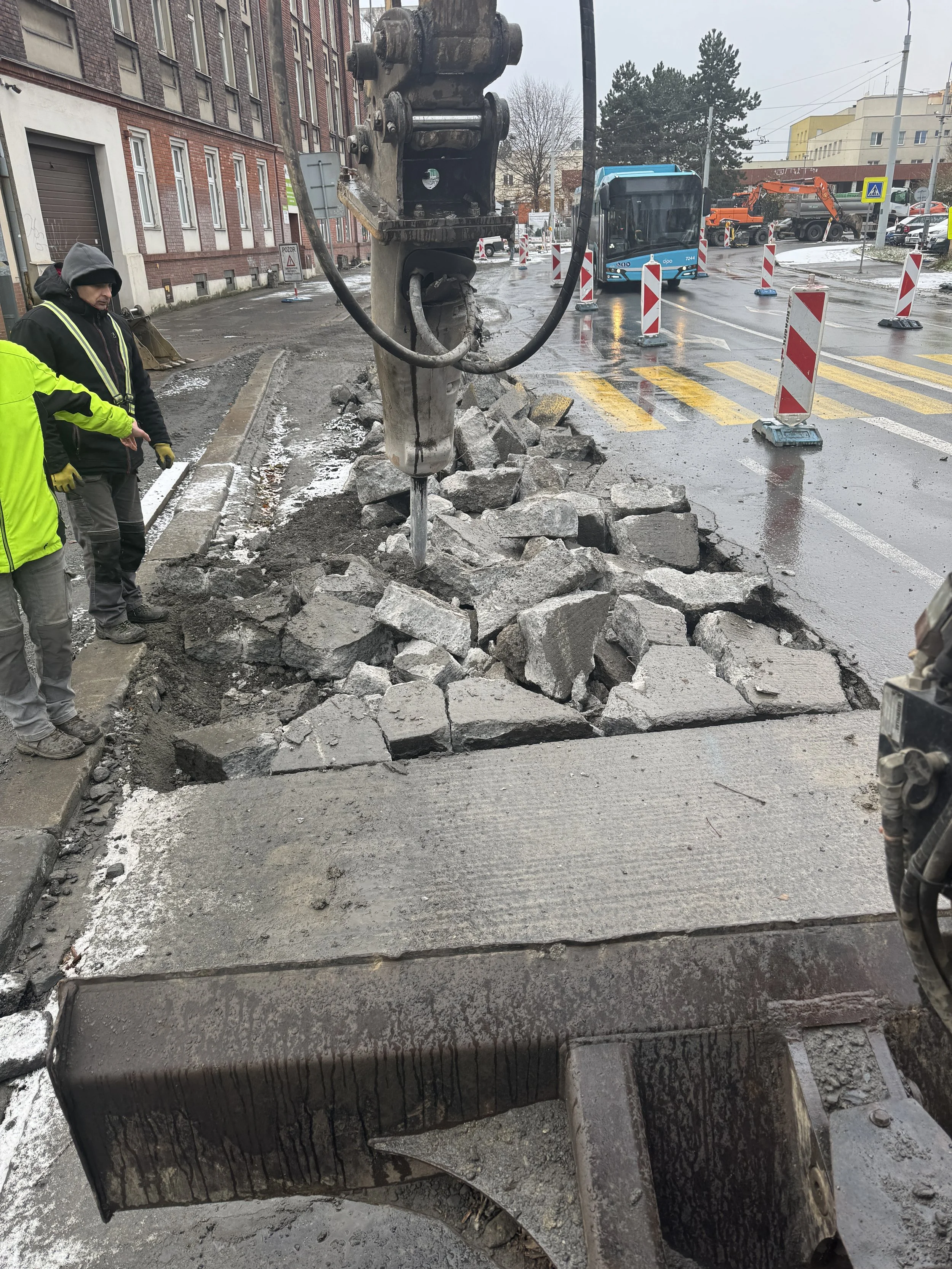 Construction workers and a large drilling machine breaking up and removing sections of an old sidewalk, with traffic cones and construction equipment in the background on a wet, overcast city street.