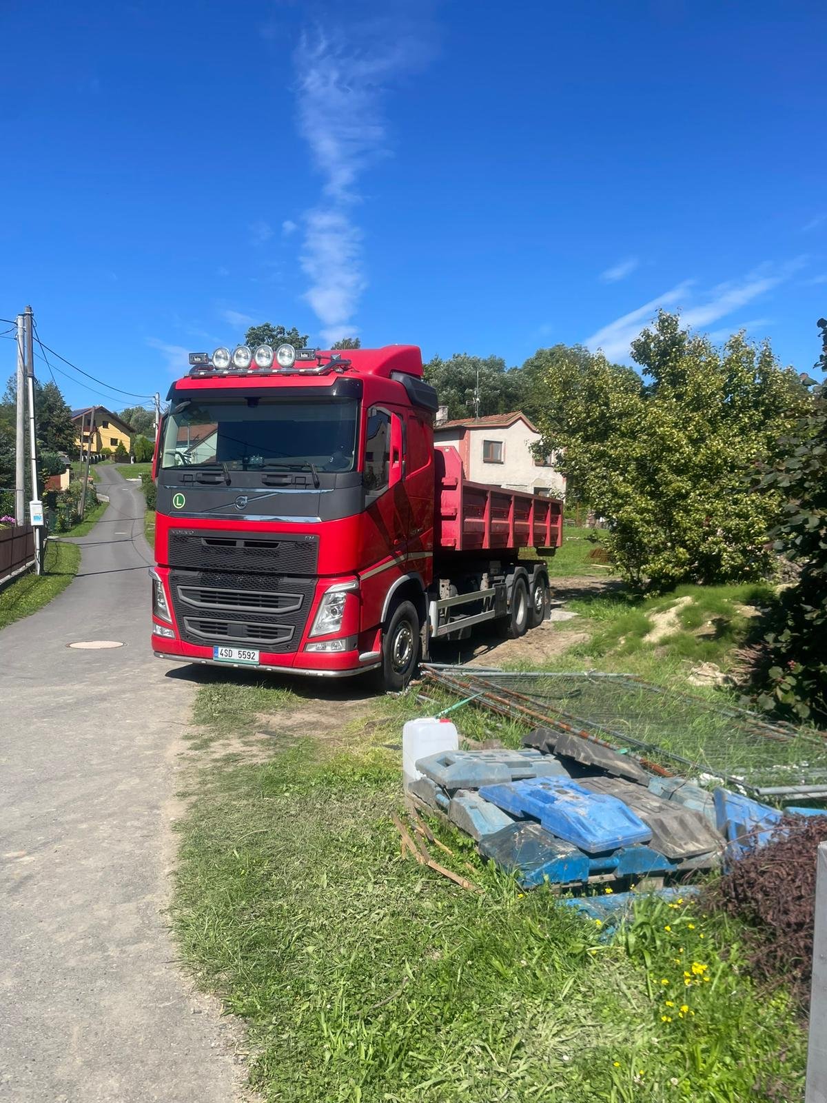 A red cargo truck parked on a grassy area beside a small dirt road in a residential neighborhood, with houses and trees in the background, under a clear blue sky.