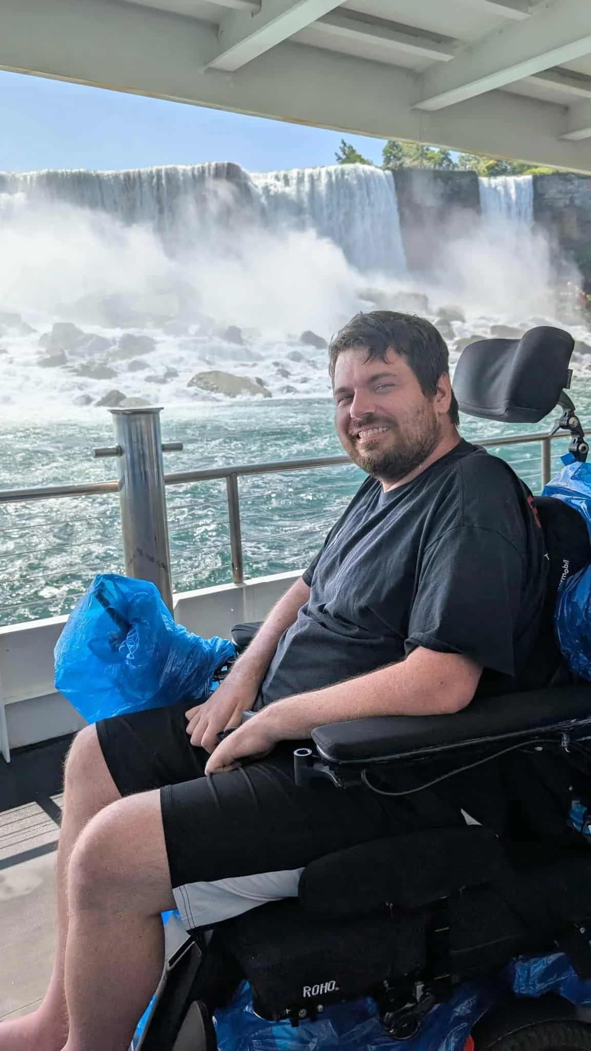 Man with a beard smiling, sitting in a wheelchair on a boat tour near Niagara Falls, with the waterfall cascading behind him.