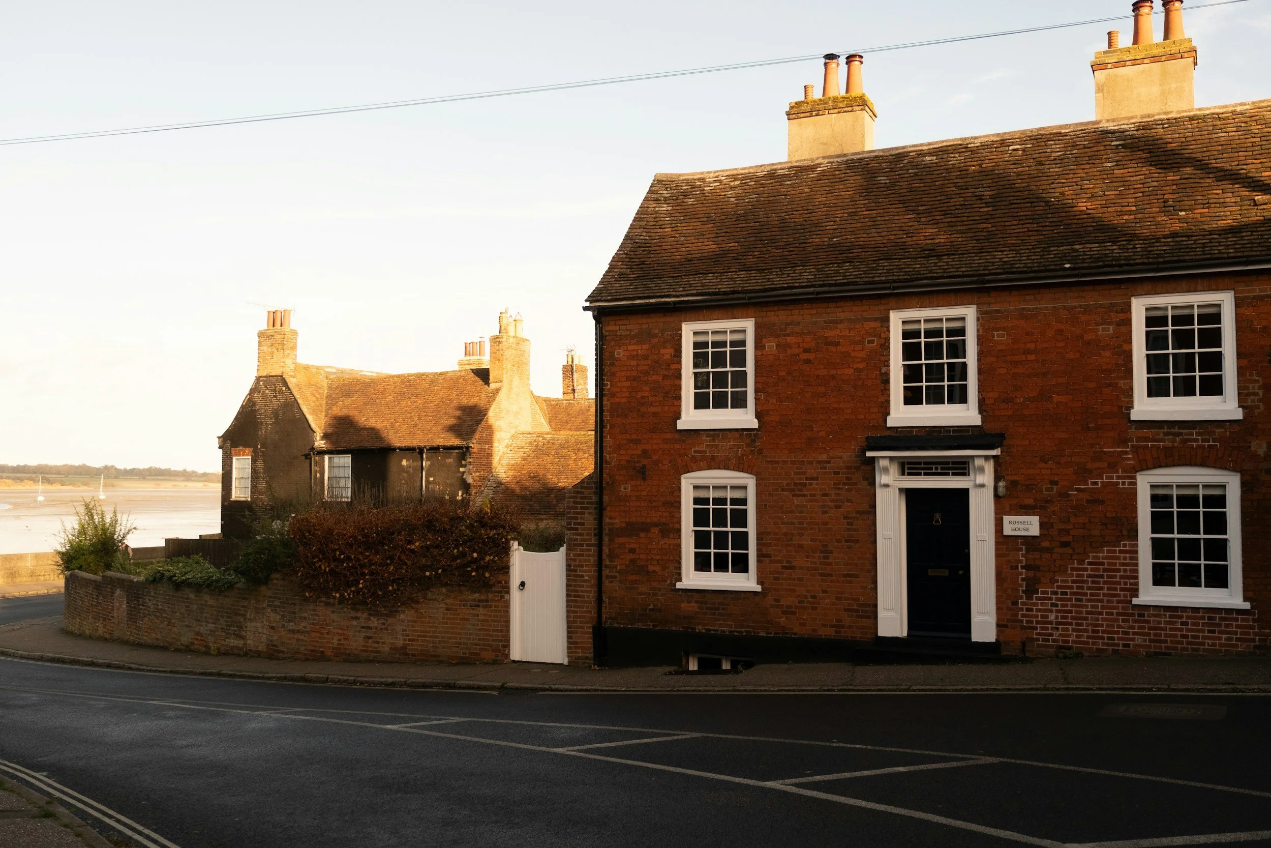 Brick house with white window frames and a black front door on a street, with additional houses and water in the background, during sunset.