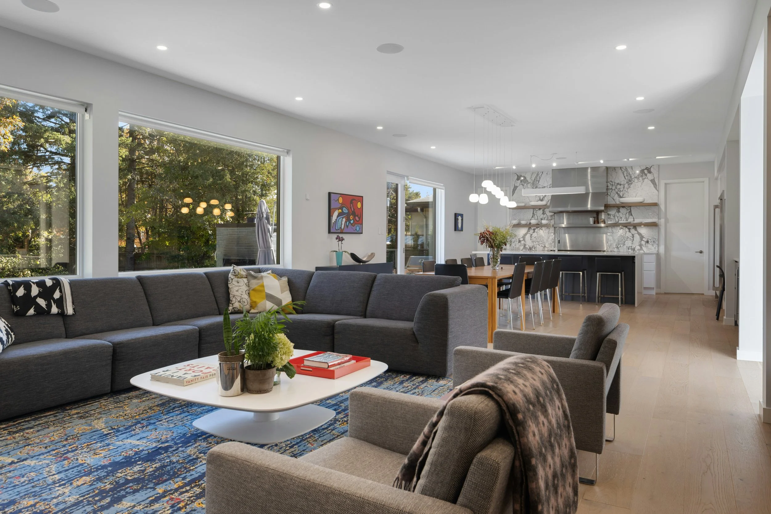 Open-concept living and dining area with large windows, a gray sectional sofa, armchair, coffee table with plants and books, dining table with black chairs, and a modern kitchen with marble backsplash and stainless steel appliances.