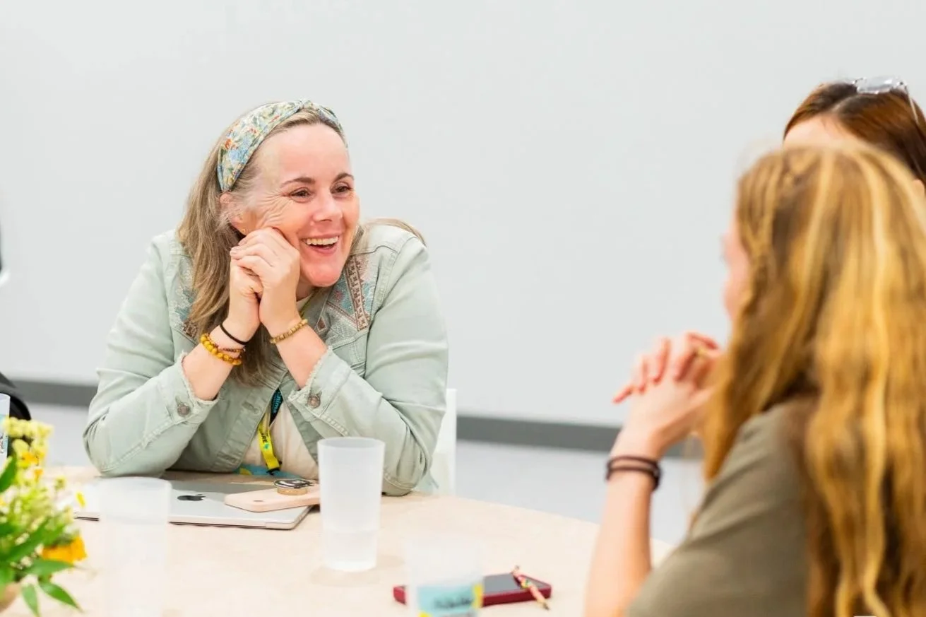 Three women sitting around a table engaging in conversation, smiling and laughing, with one woman as the main focus.