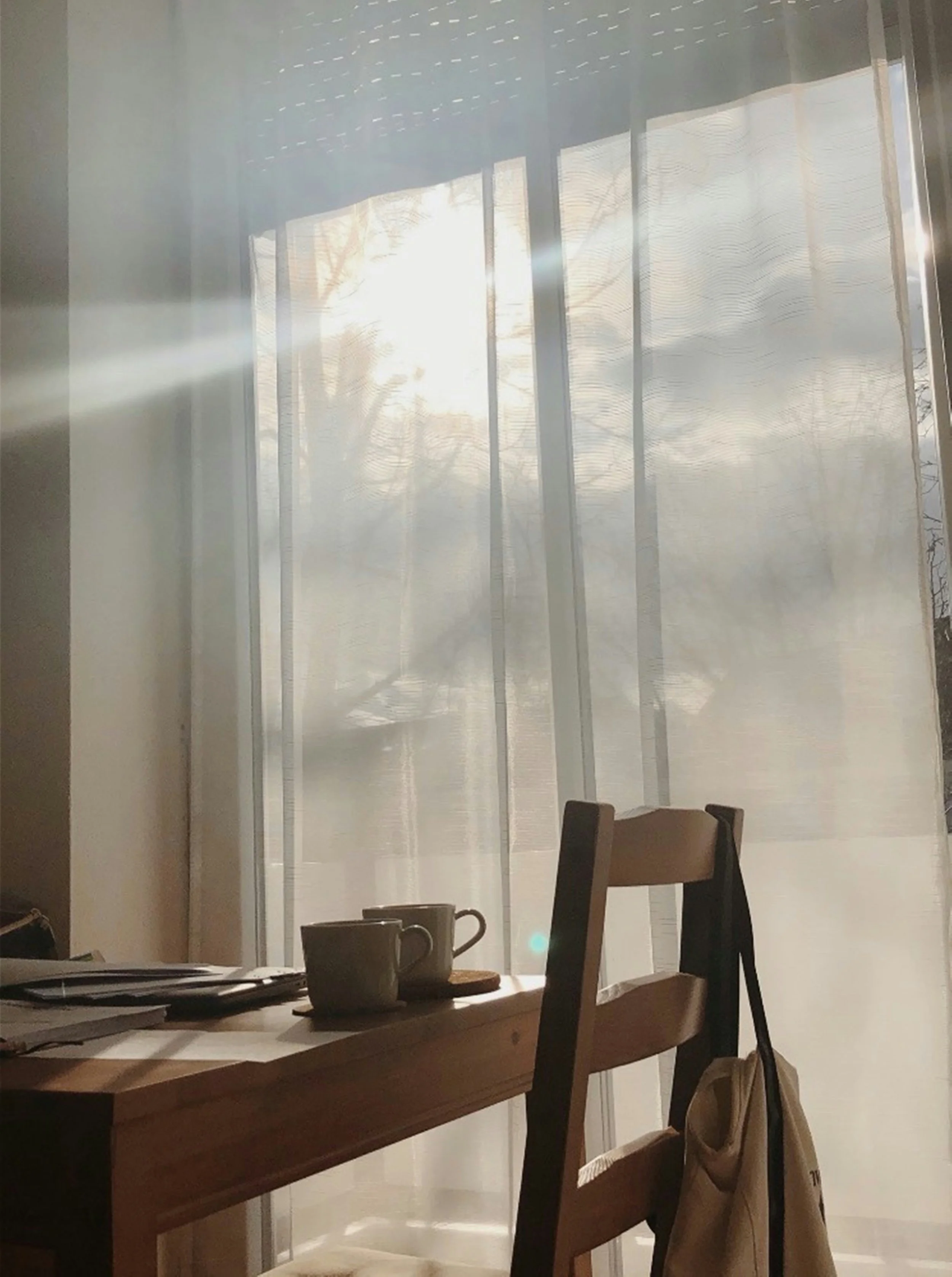 Sunlight streams through sheer curtains onto a wooden dining table with two cups and some papers, with a backpack hanging on the back of a chair.