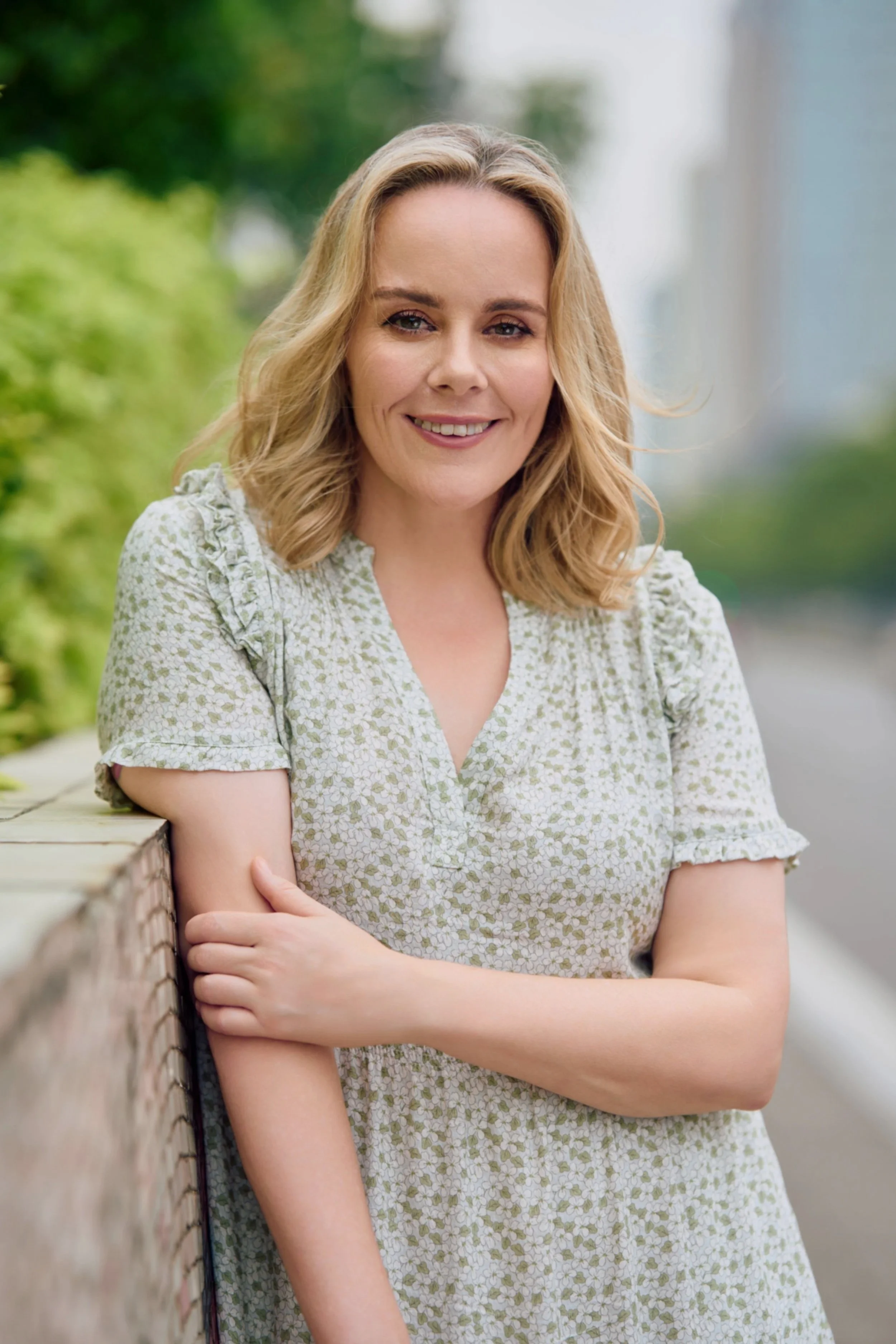 A woman smiling outdoors next to a brick wall with greenery in the background.