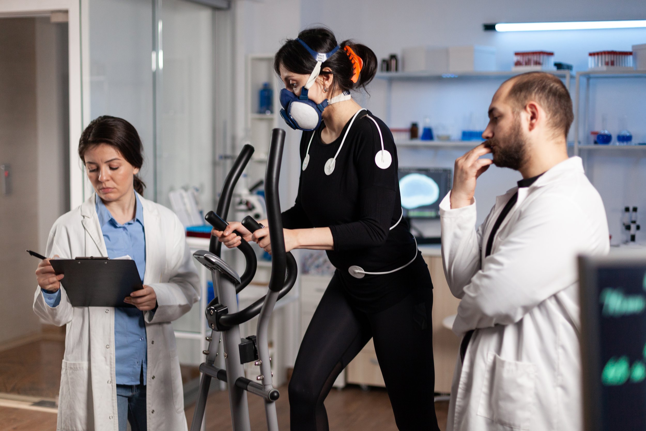 Scientists and a researcher in a laboratory with medical equipment and monitors, one woman on an exercise machine wearing a mask
