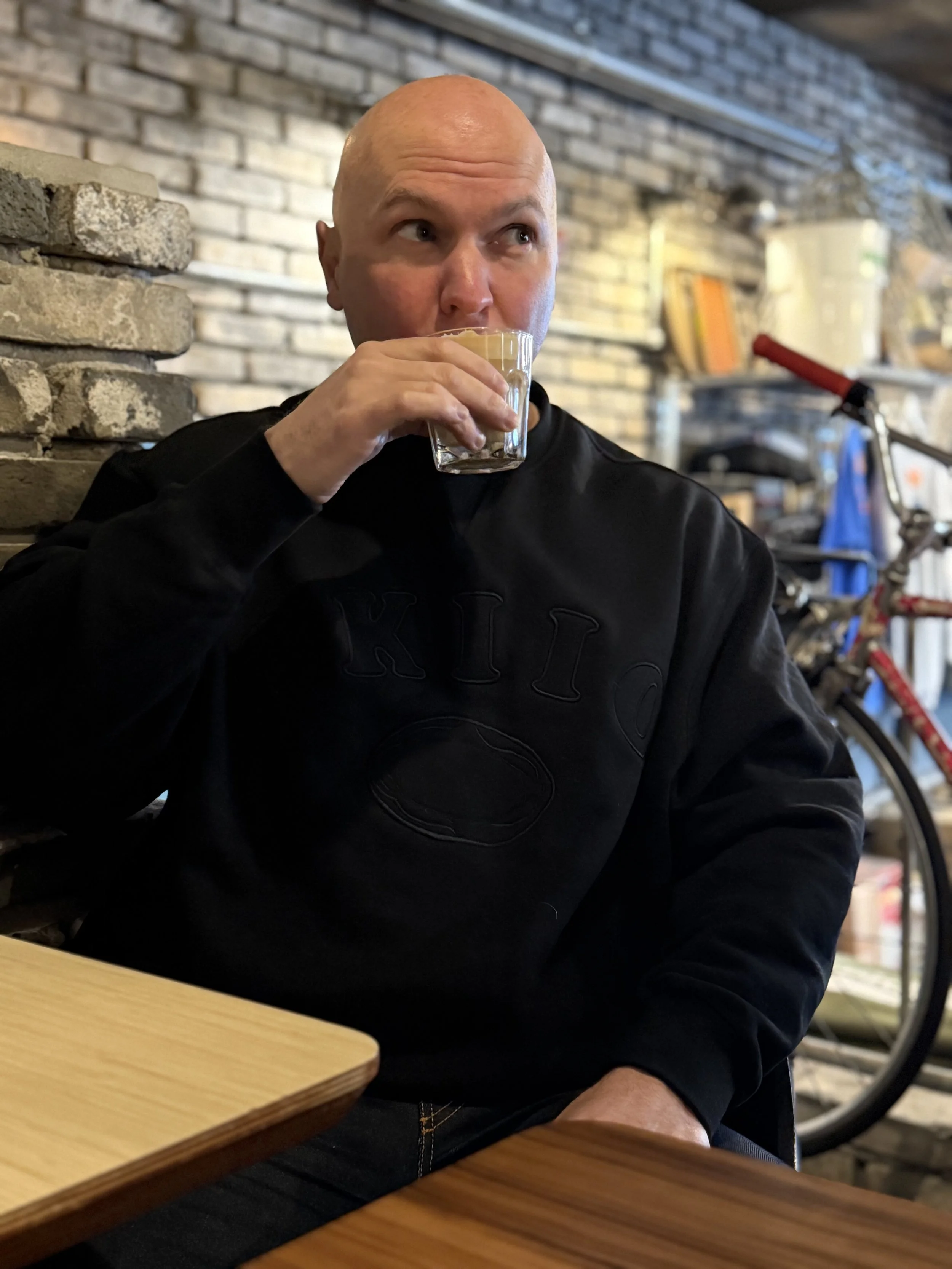 A bald man in a black sweatshirt is sitting at a wooden table, drinking a clear glass of beverage, in a cafe with a brick wall background and a bicycle in the background.