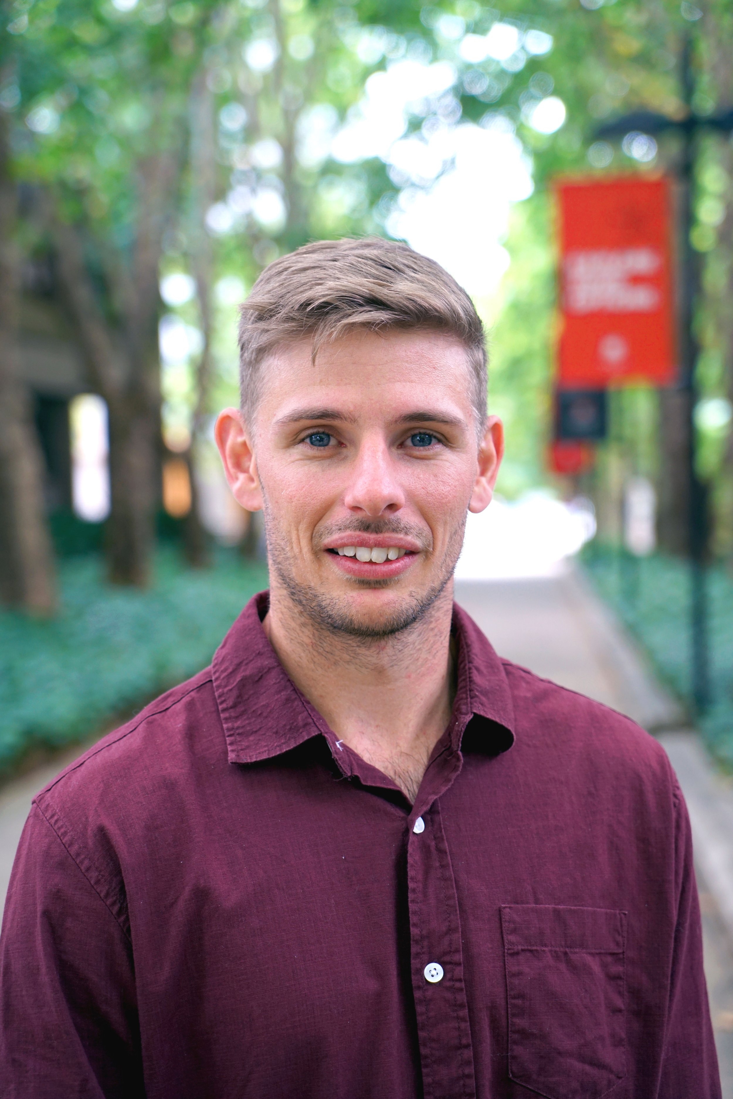 A young man with light brown hair and blue eyes, wearing a maroon button-up shirt, smiling outdoors on a tree-lined sidewalk.