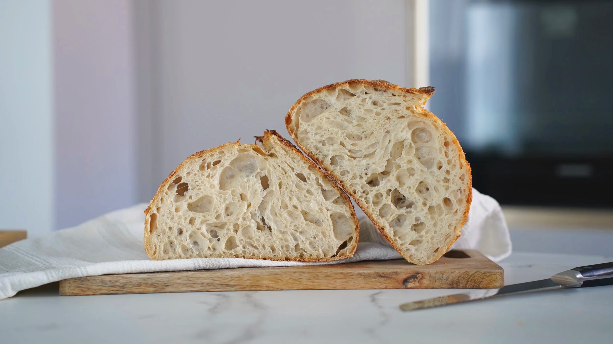 Two slices of sourdough bread on a wooden cutting board, resting on a white cloth, with a knife nearby on a white surface.