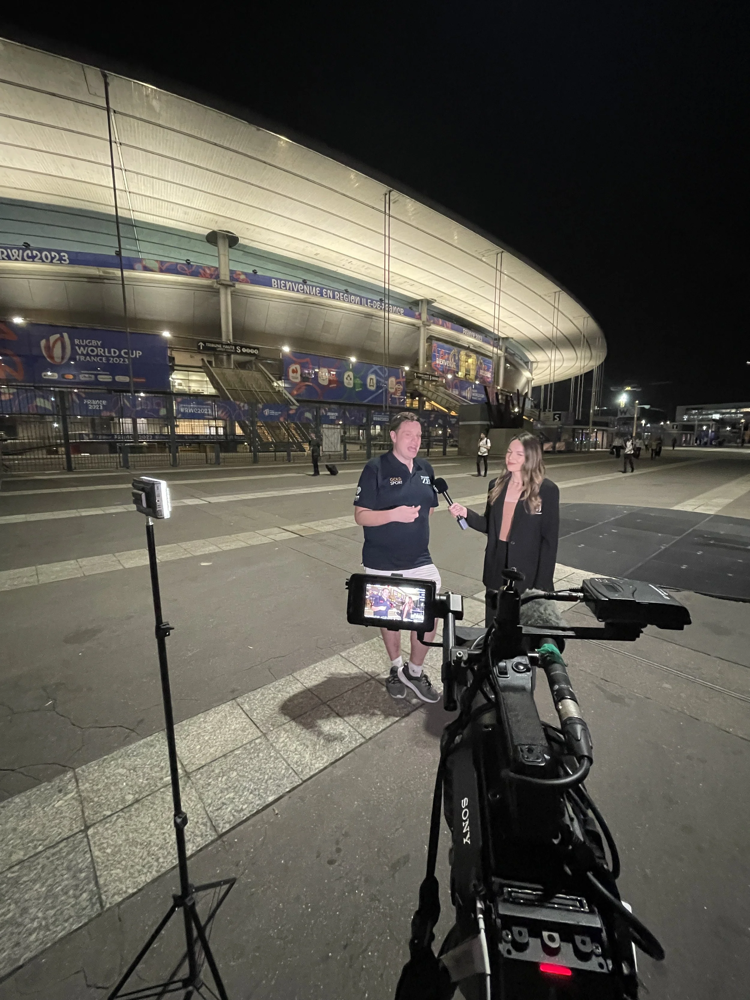 Nighttime scene outside a stadium with a man and woman being interviewed on camera. The stadium has a large, illuminated curved roof with banners related to the Rugby World Cup France 2023. Several people are visible in the background walking around or standing near the stadium.