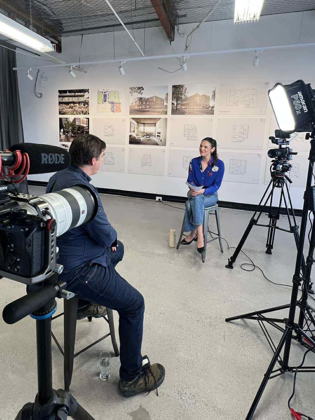 woman in blue shirt sitting on a stool being interviewed by man with camera in a studio with architectural blueprints and images on the wall