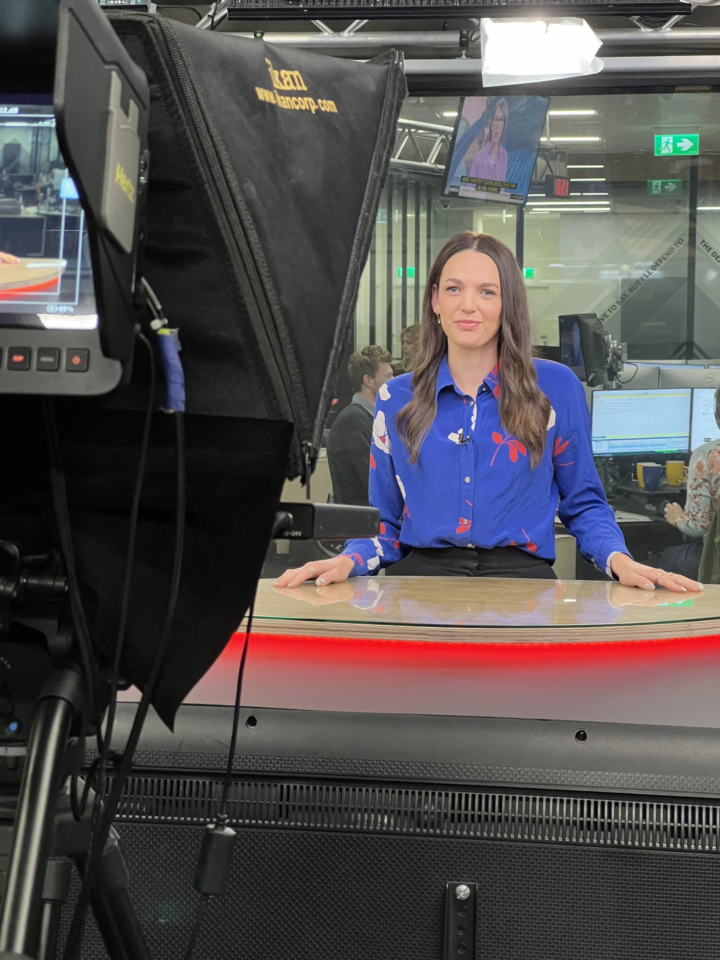 A female news anchor is being filmed at a news station, sitting at a news desk with computer monitors and news graphics on screens behind her. The camera is positioned in front of her, capturing her from the waist up.