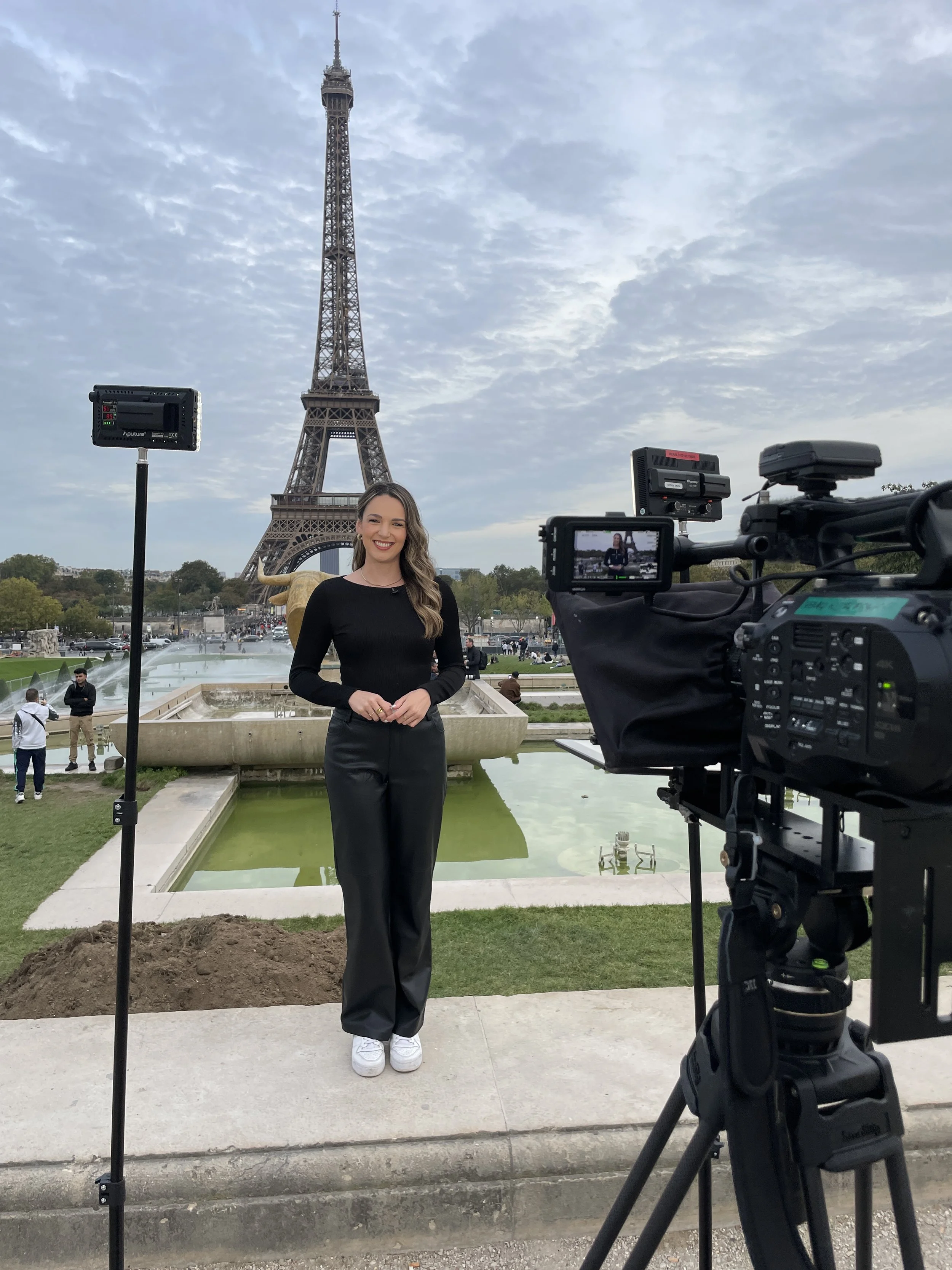 A woman in black clothing standing in front of a fountain with the Eiffel Tower in the background, during a recording setup with camera equipment and lighting.