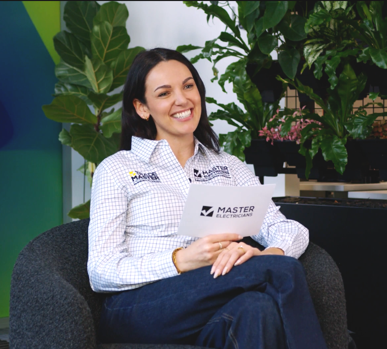 A smiling woman in a white checkered shirt with logos, sitting on a black chair surrounded by large green plants, holding a piece of paper with the Master Electricians logo.