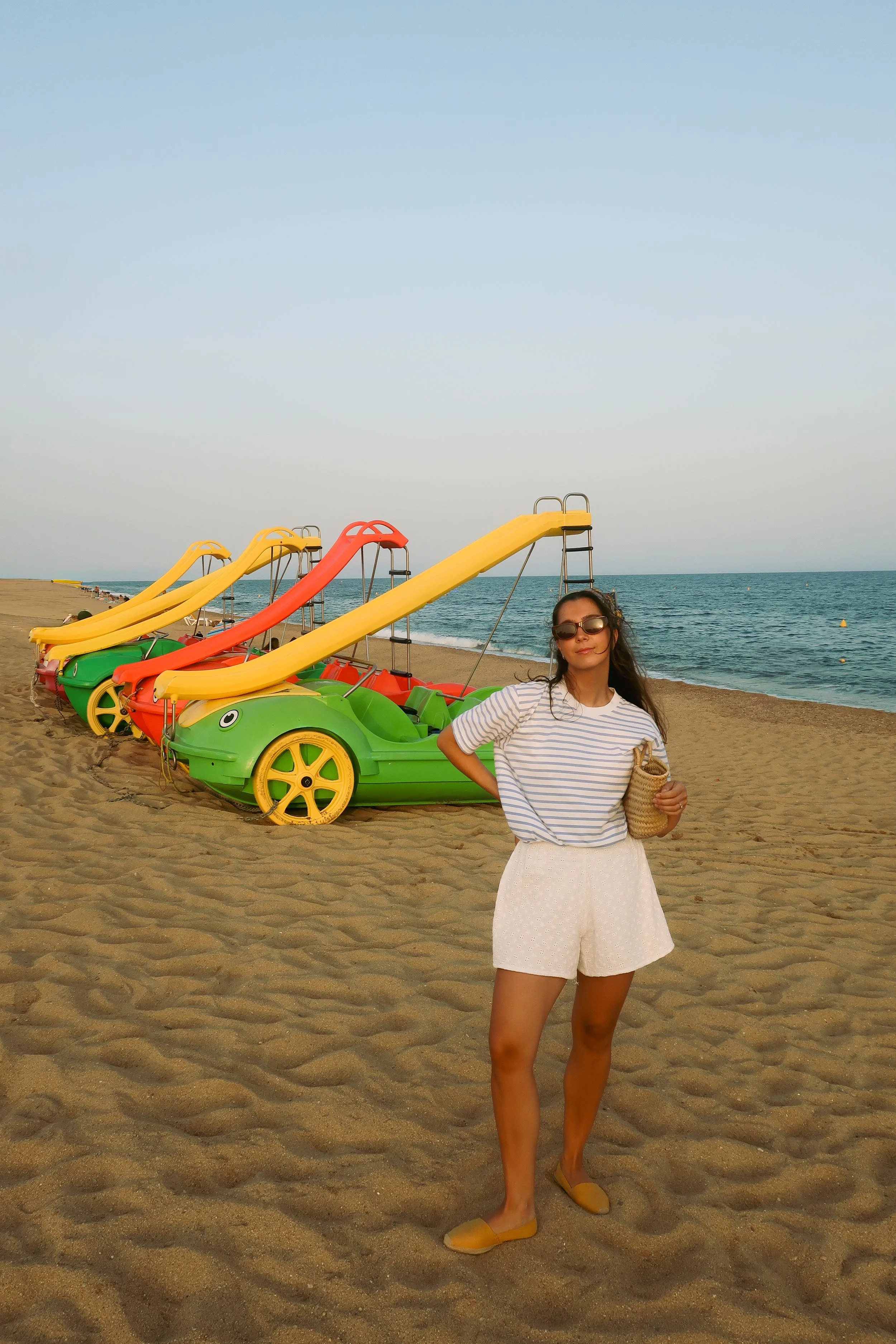A woman stands on a sandy beach posing with her hands on her hips near colorful pedal boats with slides, ocean and clear sky in the background.