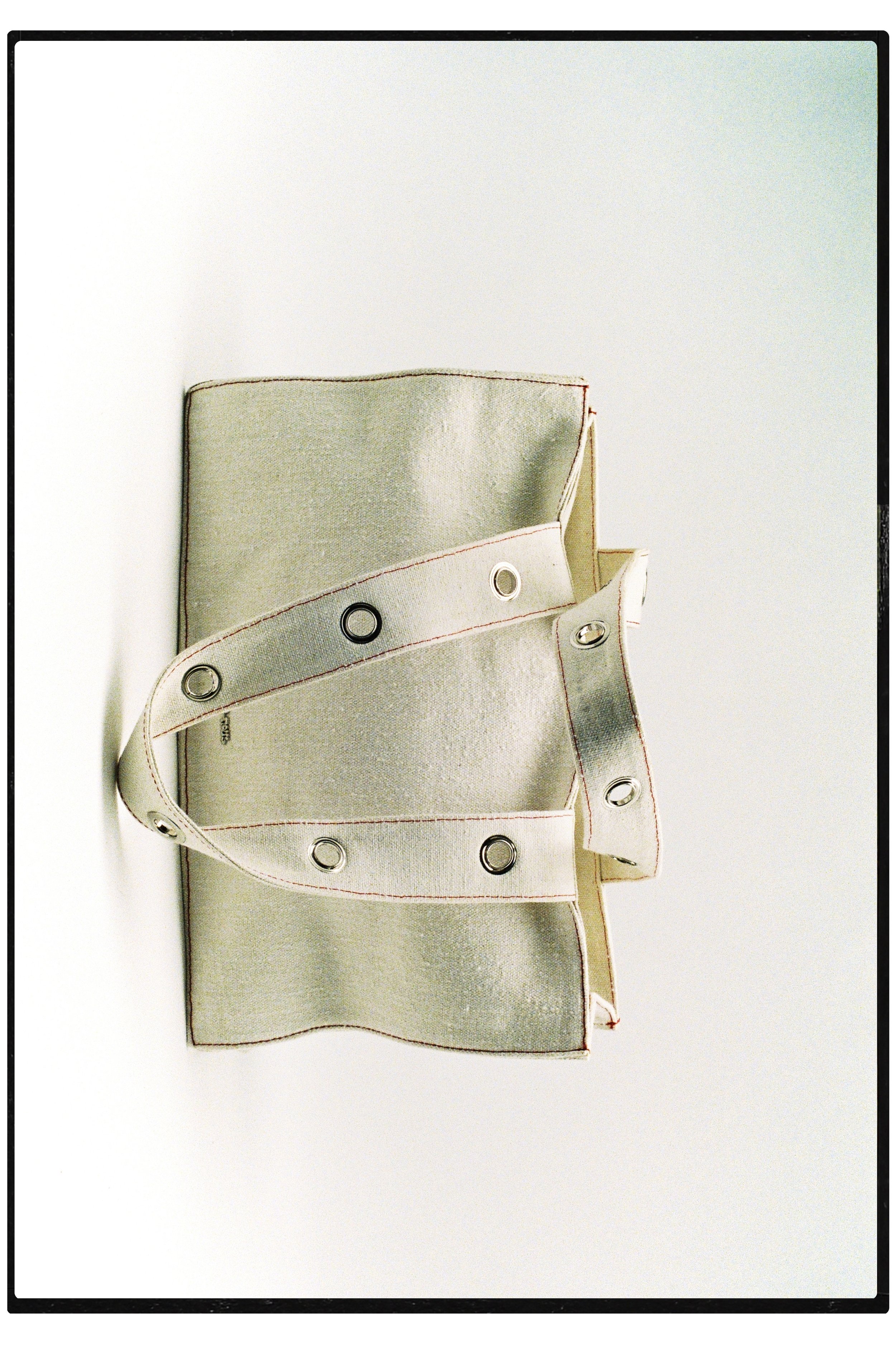 A beige fabric tote bag with metal eyelet details, lying open on a white surface.