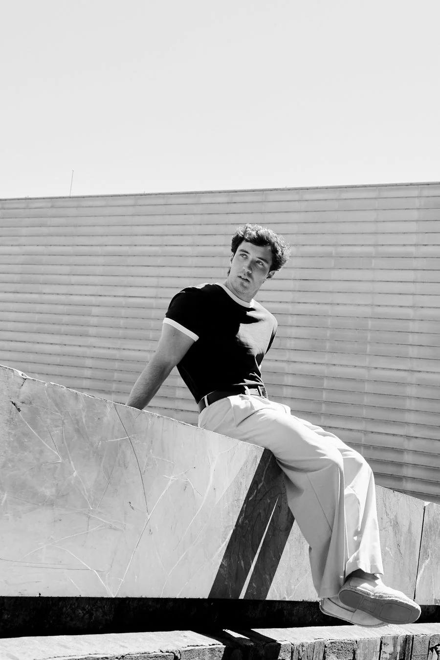 A young man sitting on a sloped marble surface outdoors, wearing a black t-shirt and wide-leg light-colored pants, with a modern building featuring horizontal siding in the background.