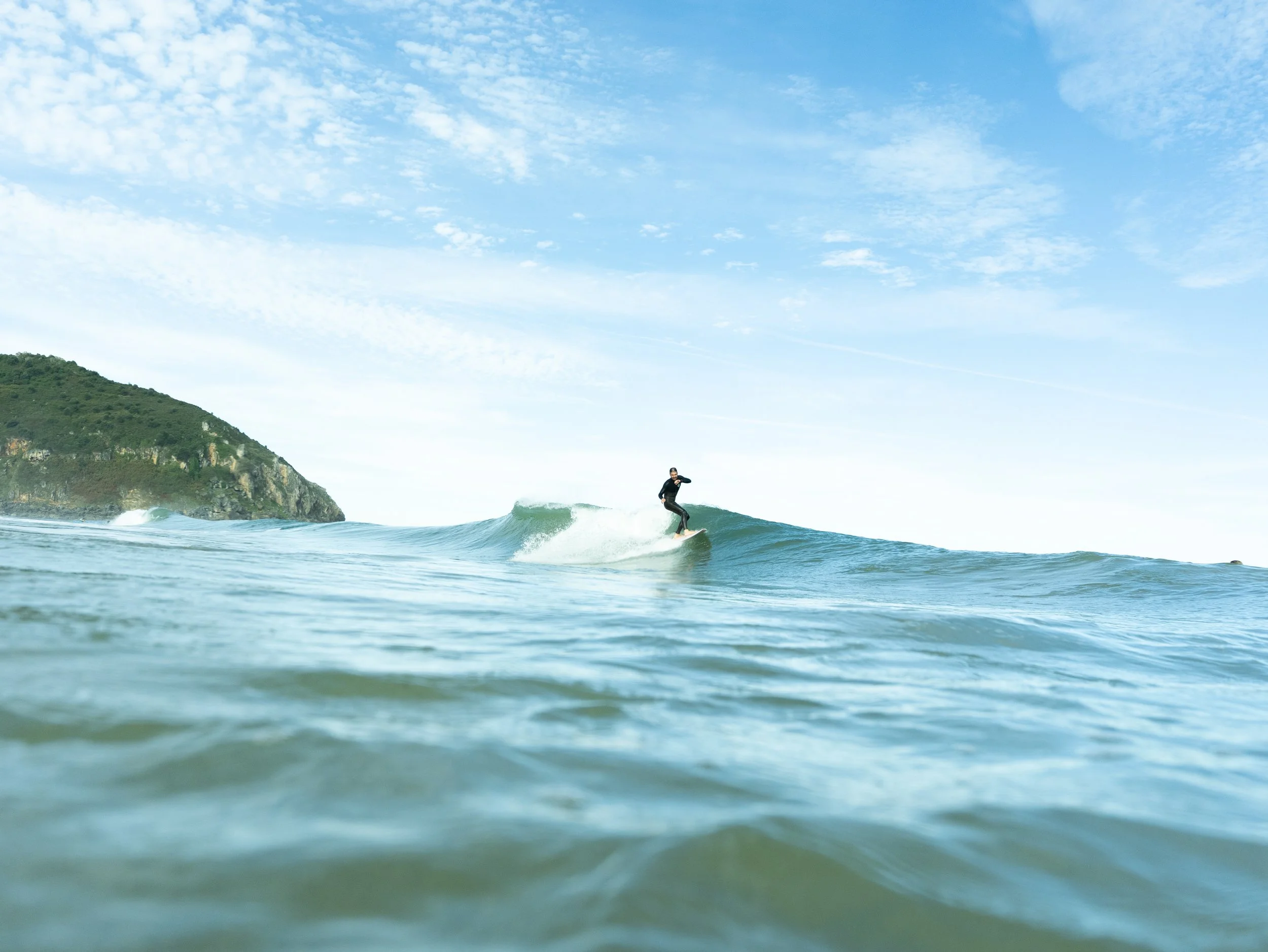 A person surfing on a wave near a coastline with a green hill and a blue sky.