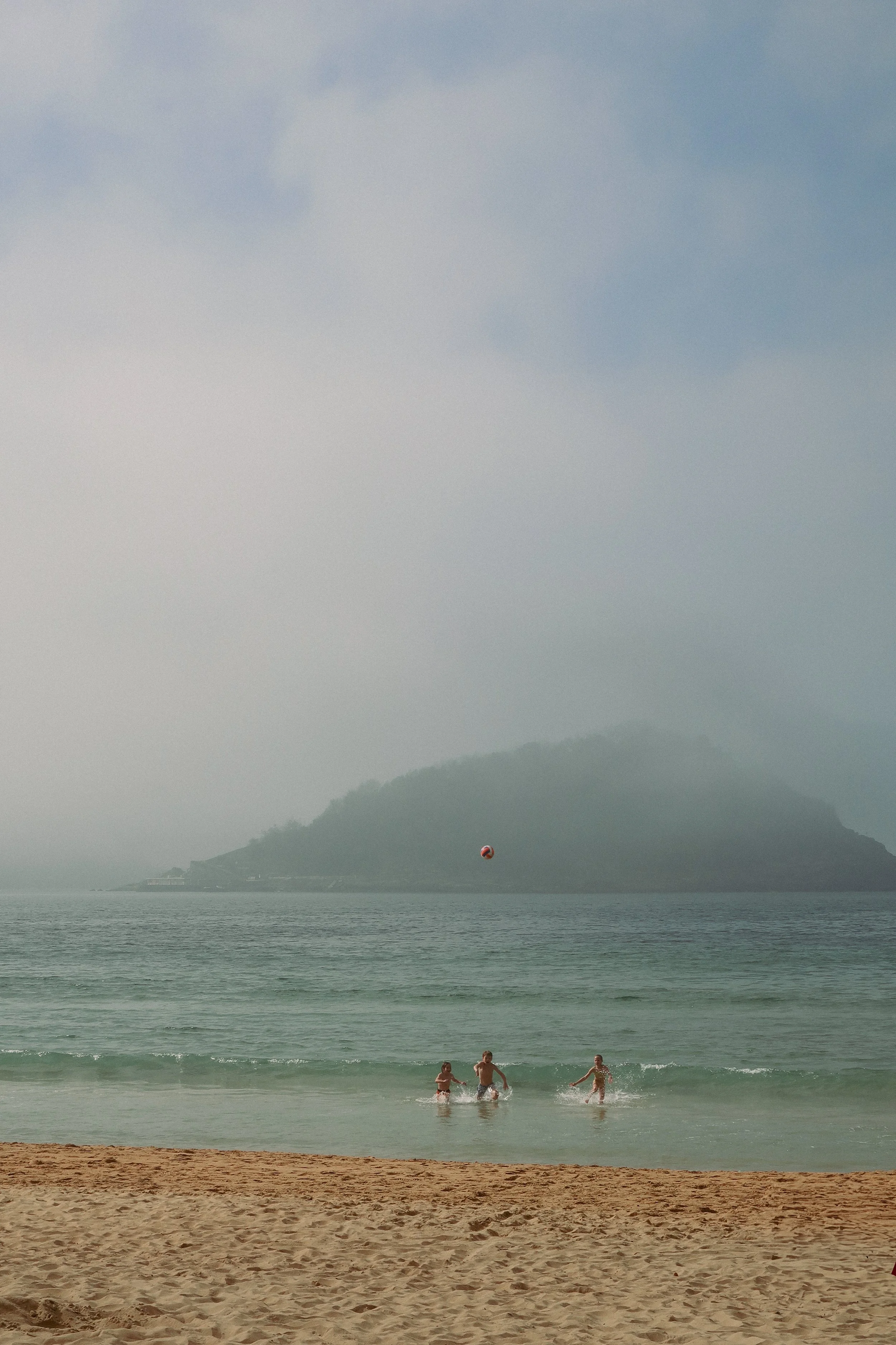 People playing in the ocean near the sandy beach with a misty island and cloudy sky in the background.