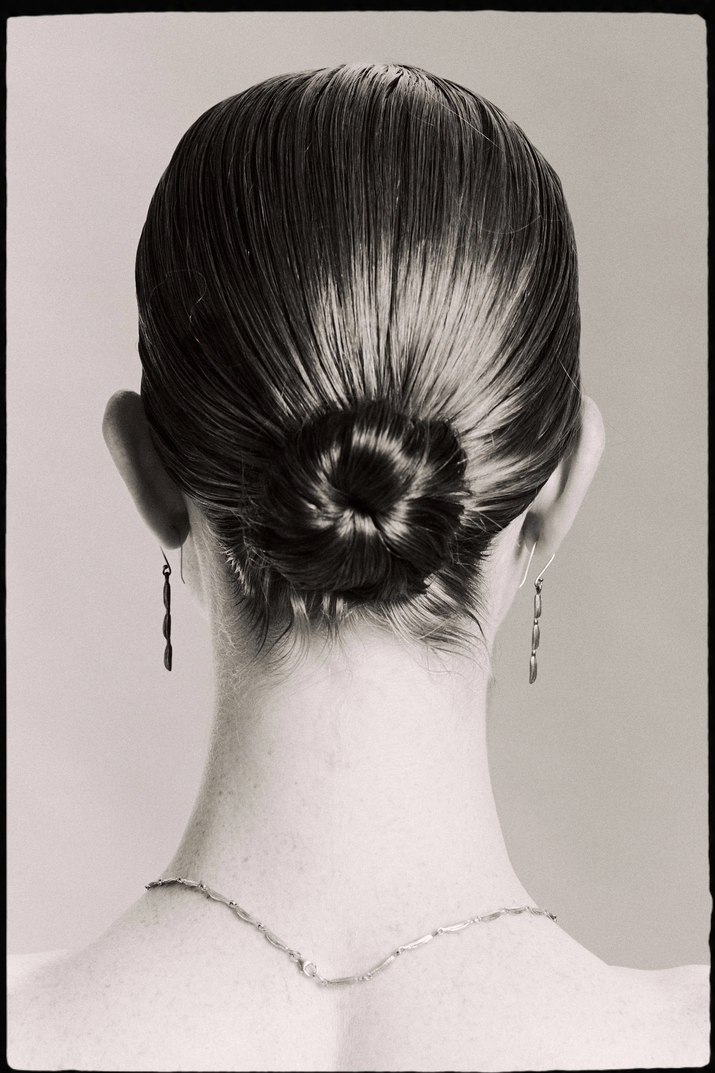 Black and white photo of a person's back, with hair styled in a bun, wearing earrings and a necklace.