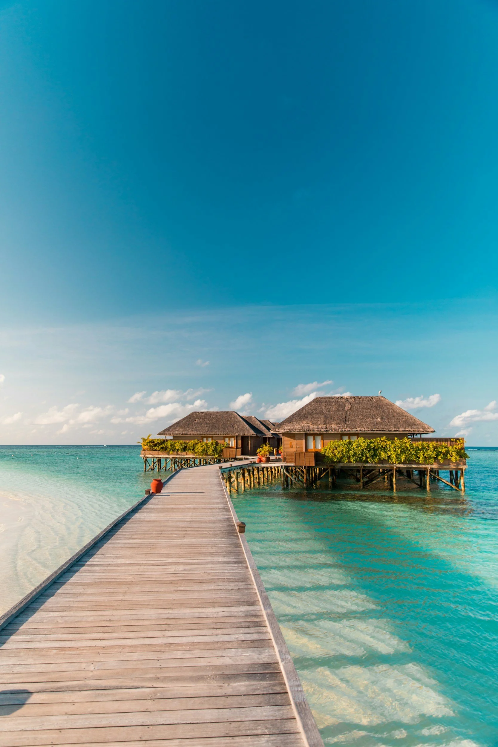 Overwater bungalows connected by a wooden pier on a clear turquoise ocean under a partly cloudy sky.