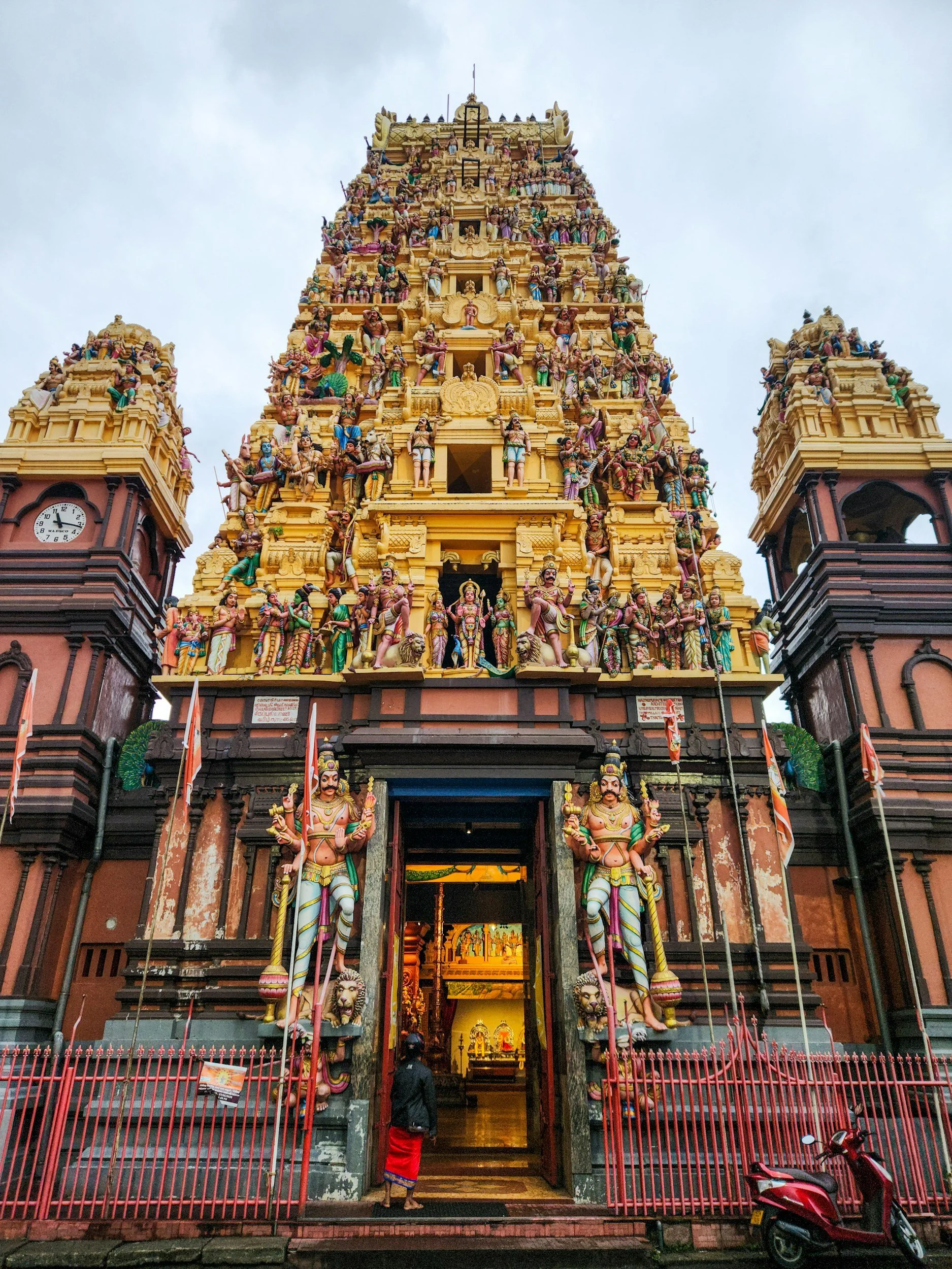 Colorful Hindu temple entrance with intricate sculptures and figures, two large guardian statues at the doorway, clock on left tower, and a person dressed in traditional attire walking inside.