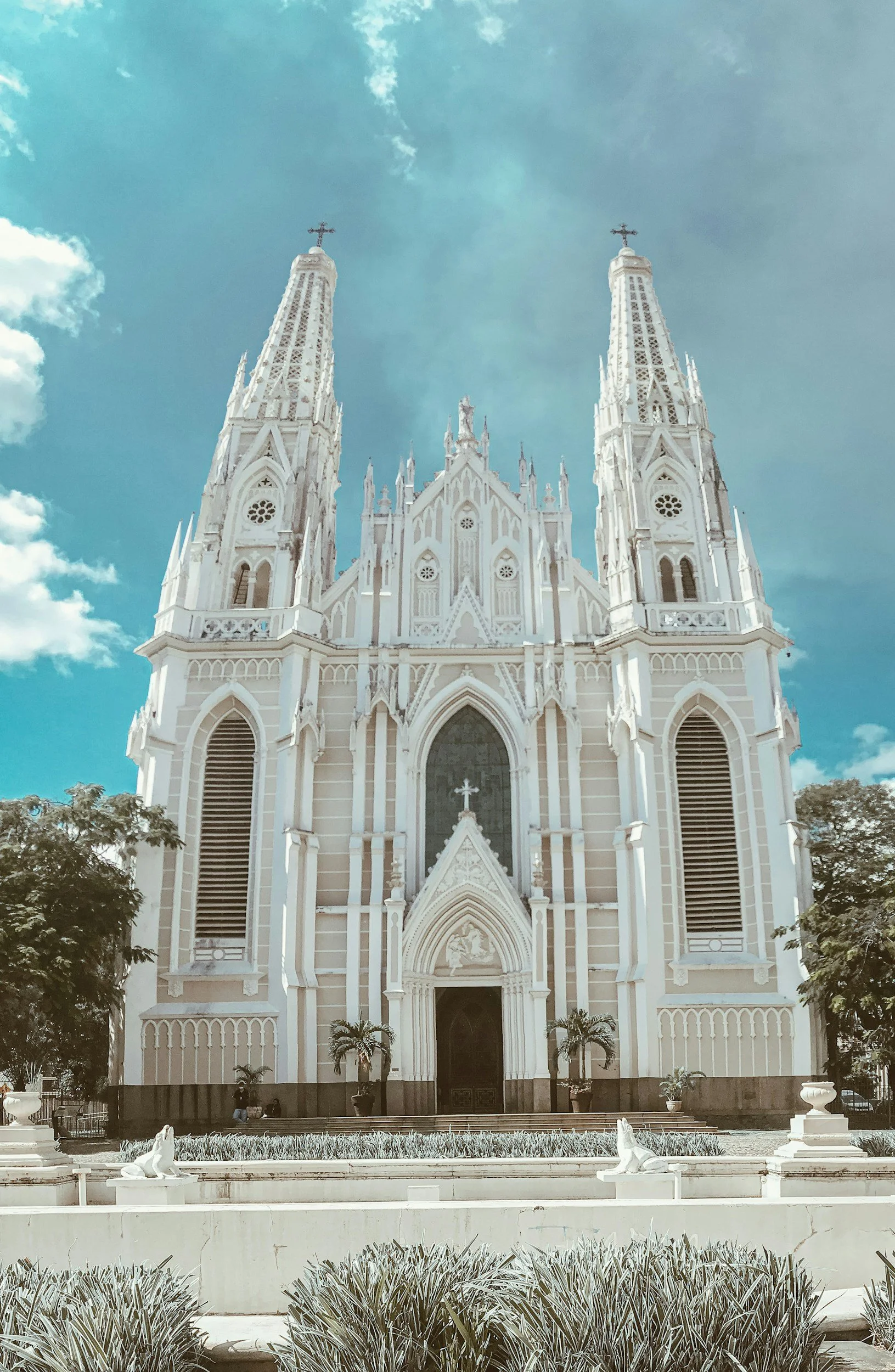 A white Gothic-style church with two tall steeples, surrounded by trees and a garden in the foreground.