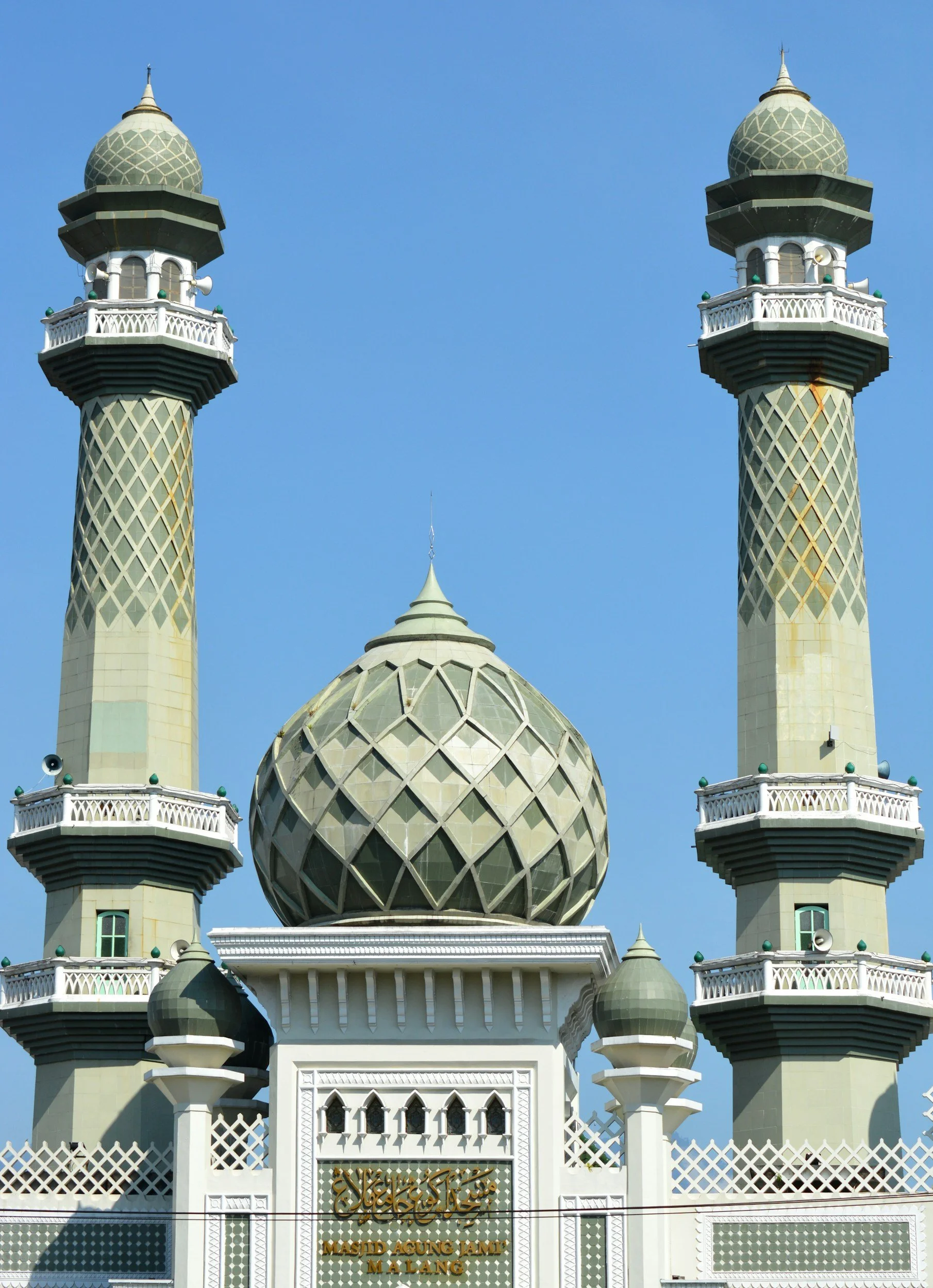 Front view of Masjid Agung Jami' Malang mosque with two minarets and a large central dome against clear blue sky.