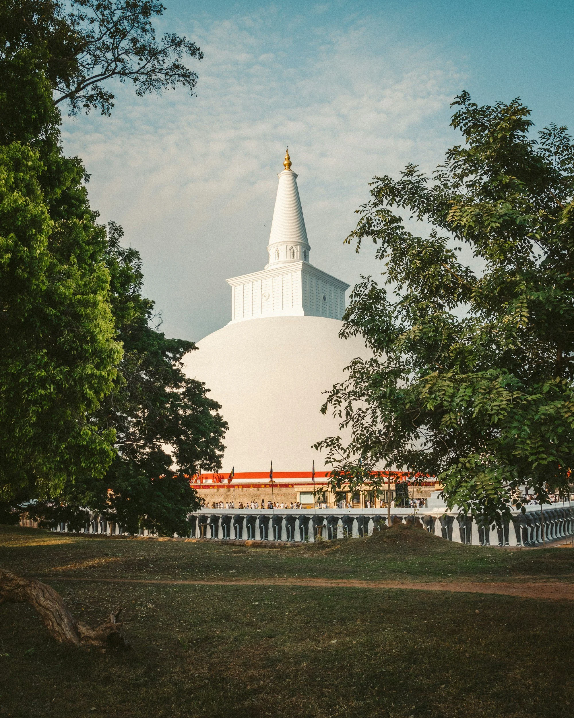 A white stupa with a tall, pointed spire surrounded by green trees and a walkway at its base