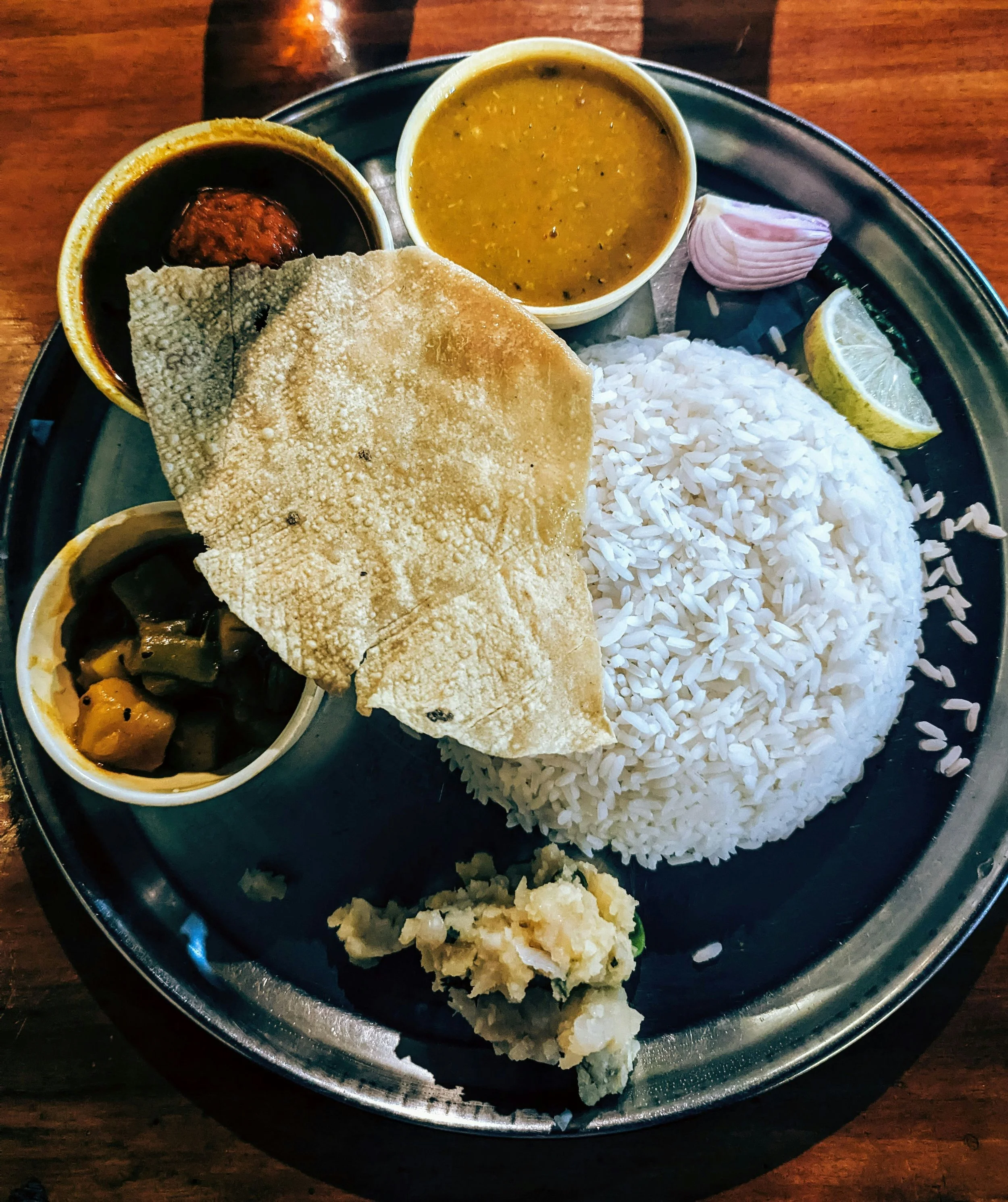 Plate of Indian cuisine with white rice, papadum, lentil curry, vegetable pickle, chickpea curry, lemon wedge, onion, and potato salad.