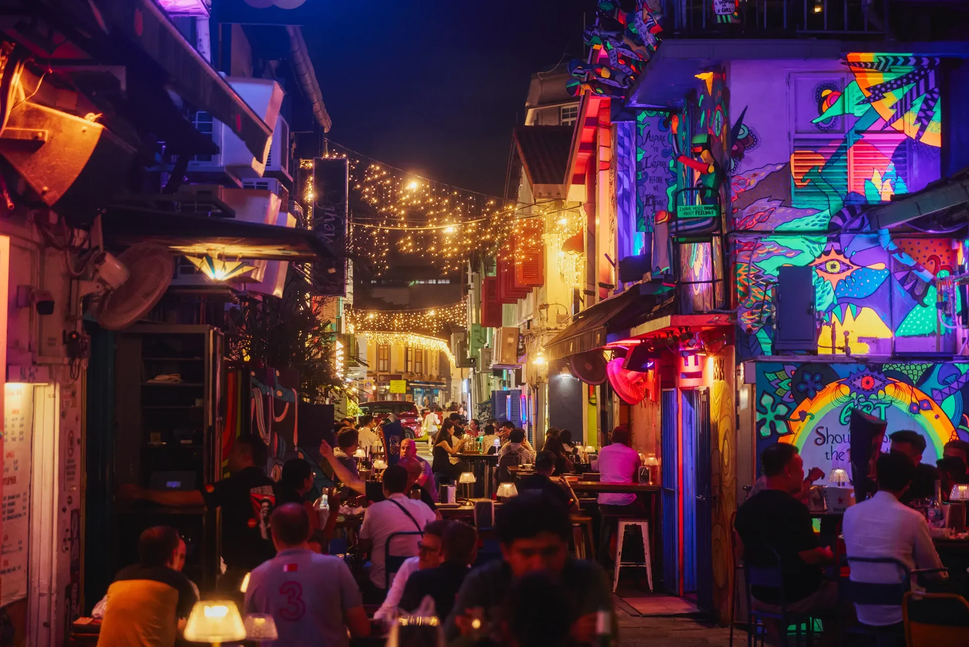 Night scene of an outdoor street with colorful graffiti art on walls, string lights overhead, and people dining at tables