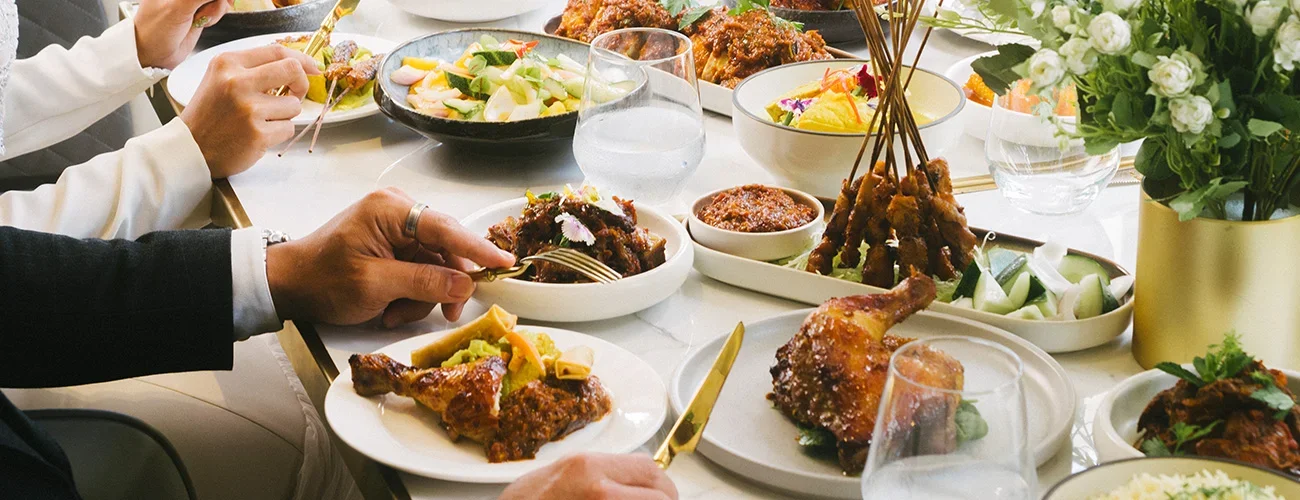 A table set with various dishes including grilled chicken, skewered meat, salad, and sauces, with people serving themselves and a vase of flowers in the background.