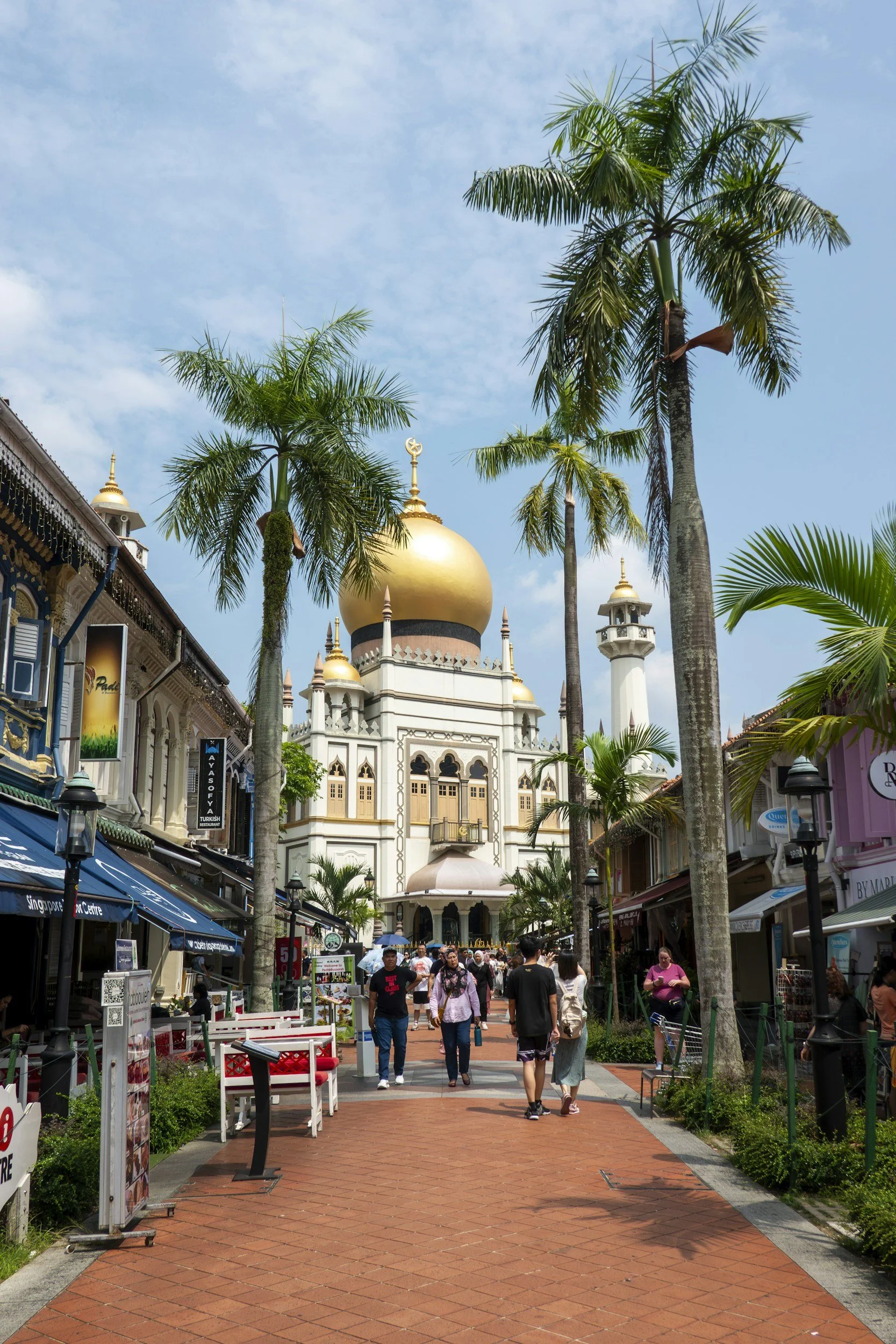People walking towards a replica of the Taj Mahal surrounded by palm trees in a lively outdoor shopping area.