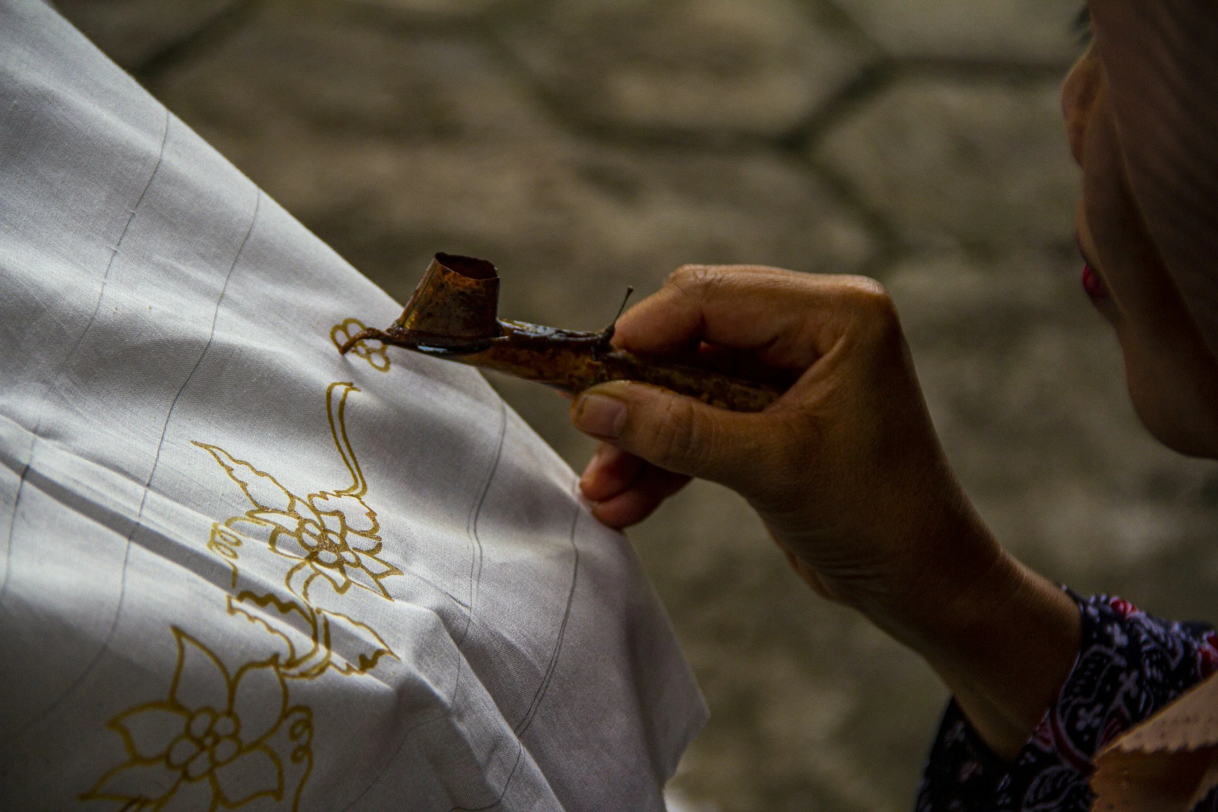 A person is sewing a golden thread onto white fabric with floral embroidery, using a thimble on their finger.