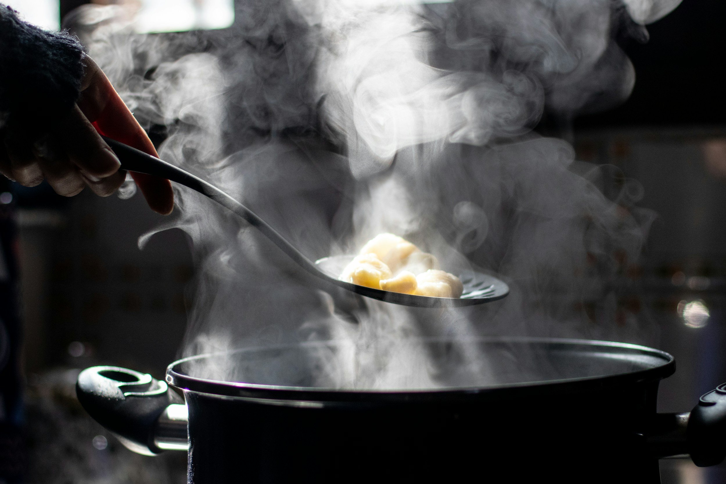 A person holding a spoon with steamed dumplings over a pot of boiling water, with steam rising in the air.